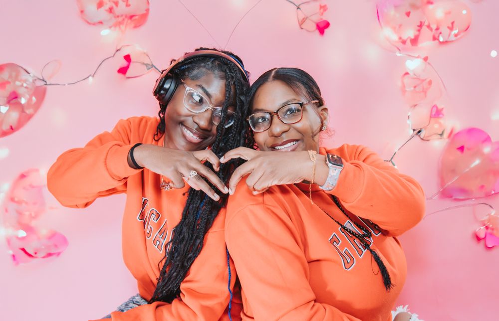 Two smiling women in matching orange sweatshirts make heart shapes with their hands in front of pink heart balloons.