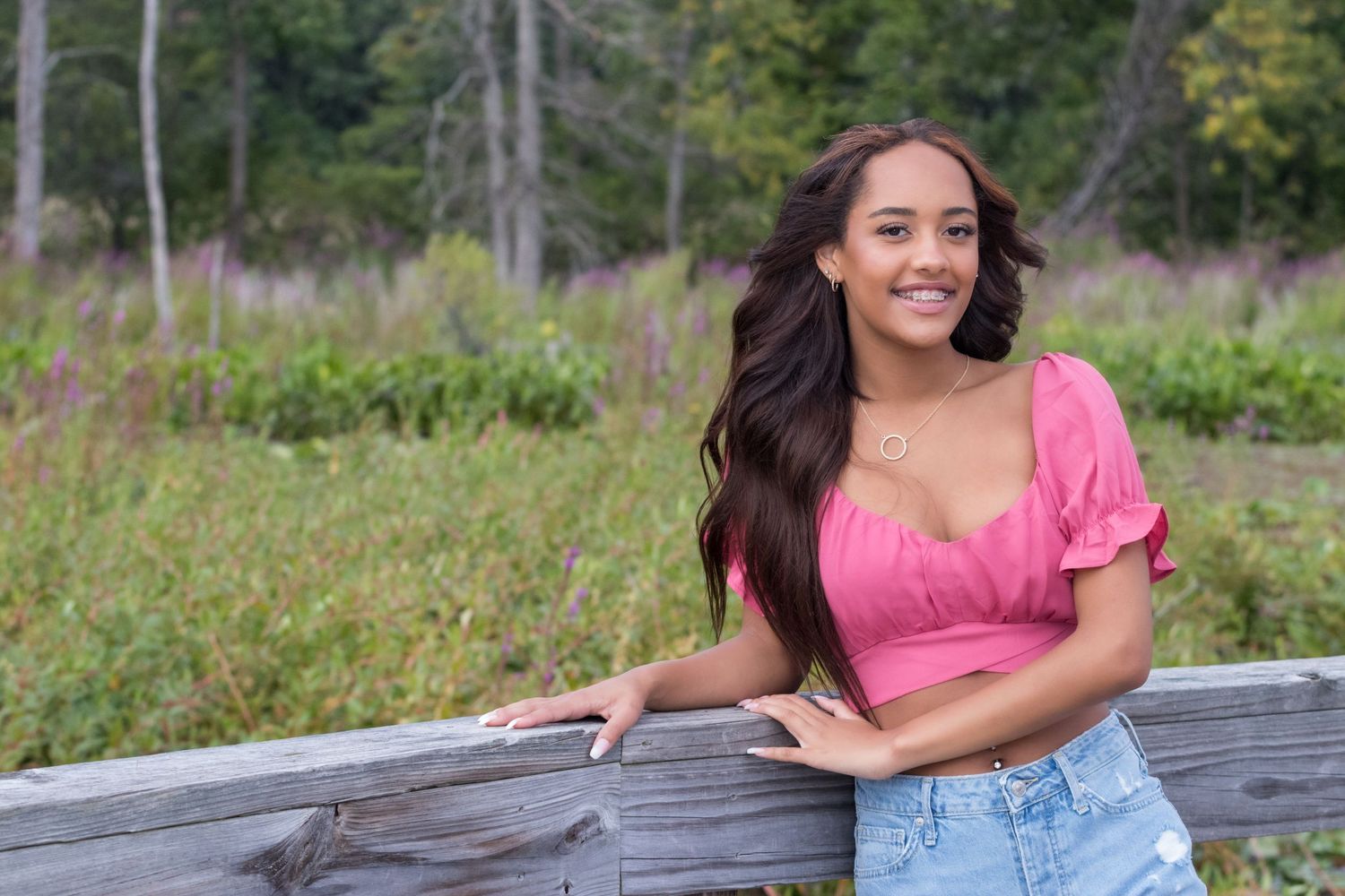 Smiling young woman in a pink top and denim shorts leans on a wooden fence with a wildflower meadow and trees behind her.