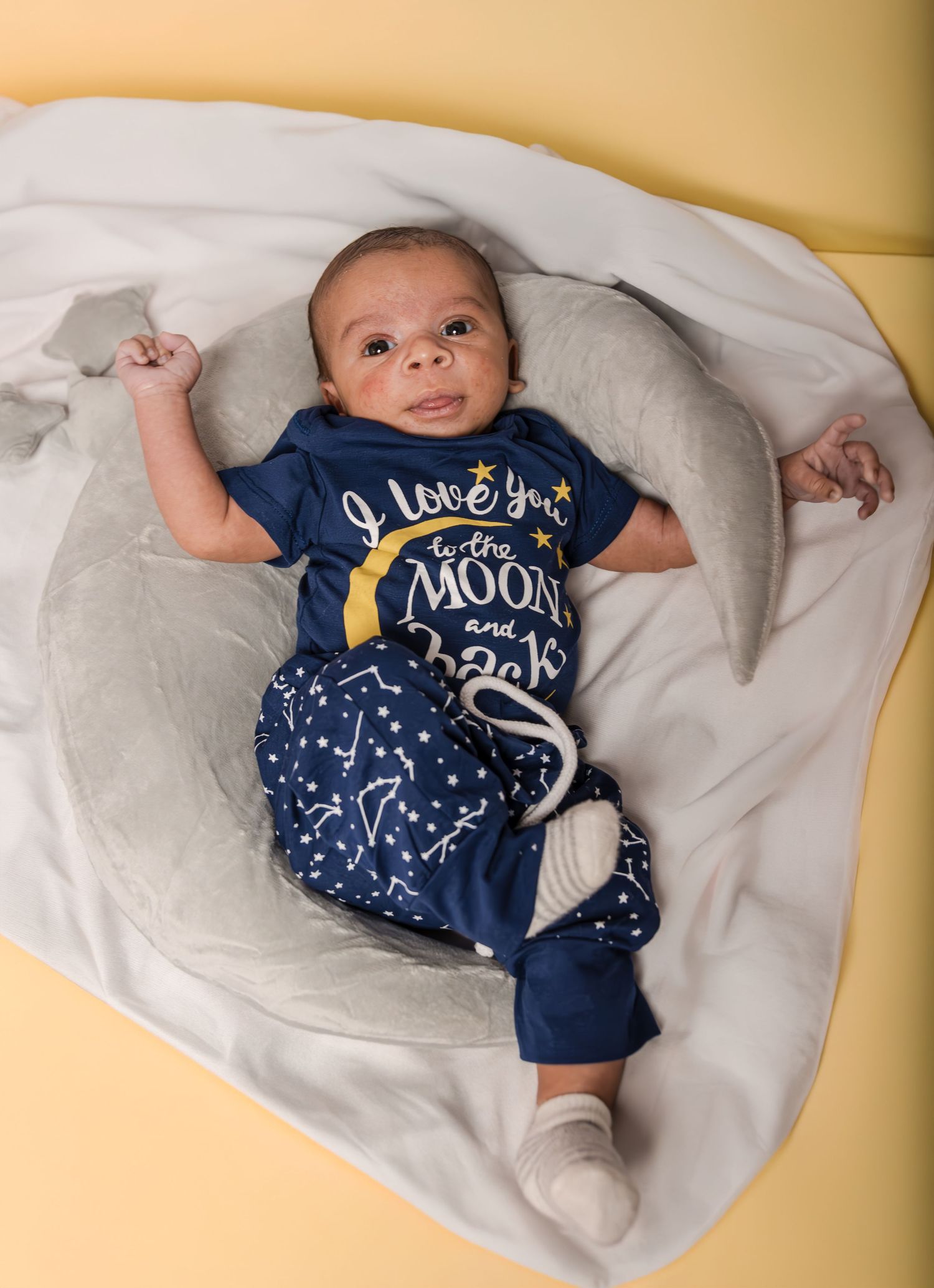 A smiling baby in a navy moon-and-stars outfit lies on a gray moon-shaped pillow and white blanket against a yellow backdrop.