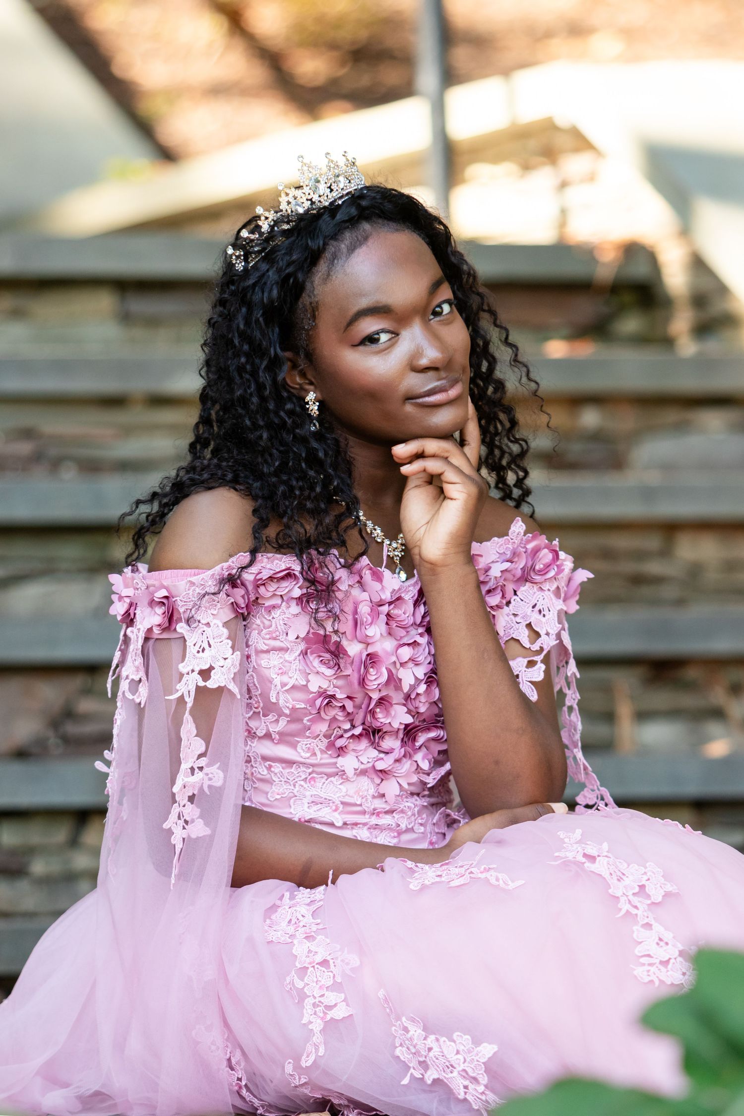 Young woman in a pink lace gown and jeweled tiara rests her chin on her hand while posing on stone steps.