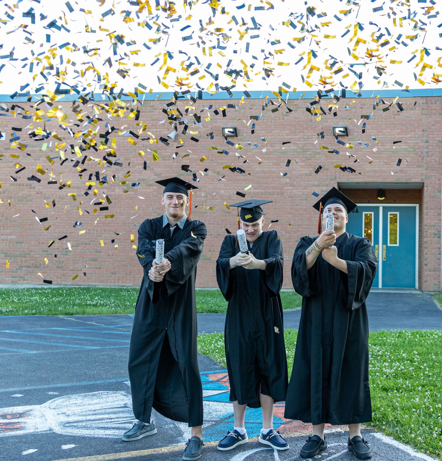Three graduates in caps and gowns hold rolled diplomas while confetti showers down in front of a brick school building.