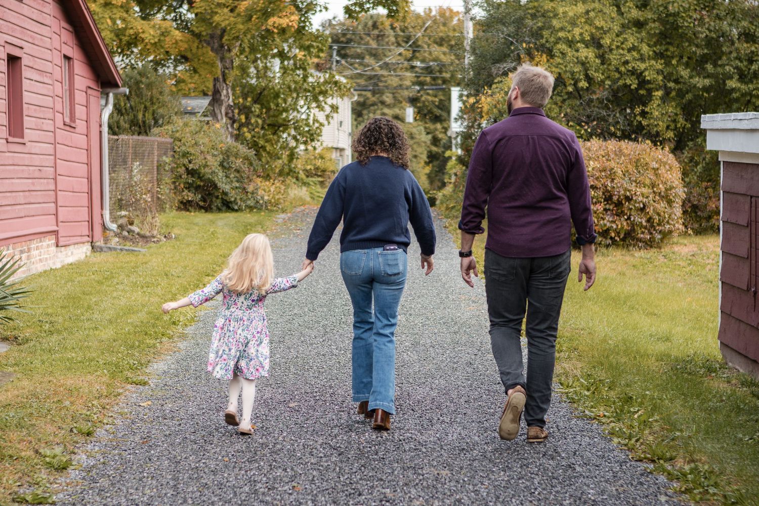 A family of three walks away on a gravel path, with a little girl in a floral dress holding her mother's hand.