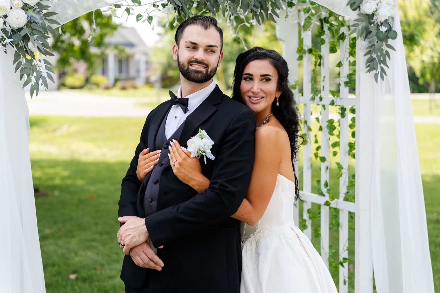 A smiling groom in a black tuxedo and bow tie embraces his bride in a white gown beneath a floral wedding arch.