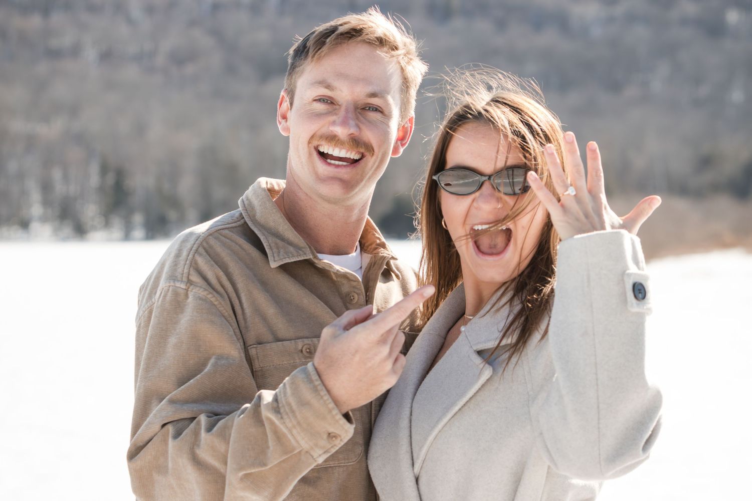 A joyful newly engaged couple laughs outdoors in the snow while the woman holds up her hand to show off her engagement ring.