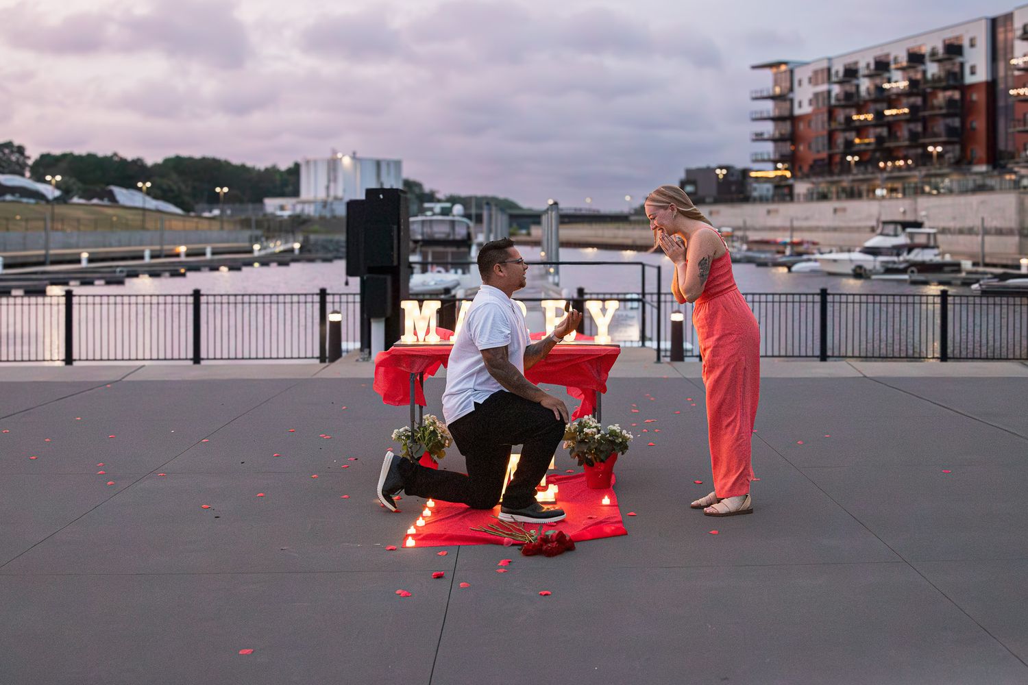 A man kneels on one knee proposing to a woman in a coral dress beside a lit marquee sign and candles on a waterfront promenade.