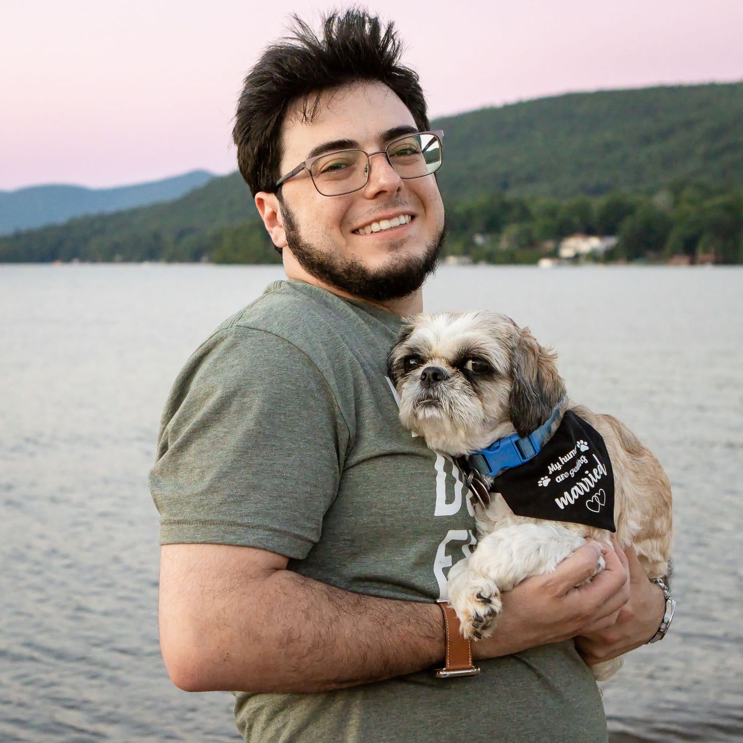 Smiling bearded man with glasses holds a small shih tzu in a bandana beside a lake with hills at sunset.