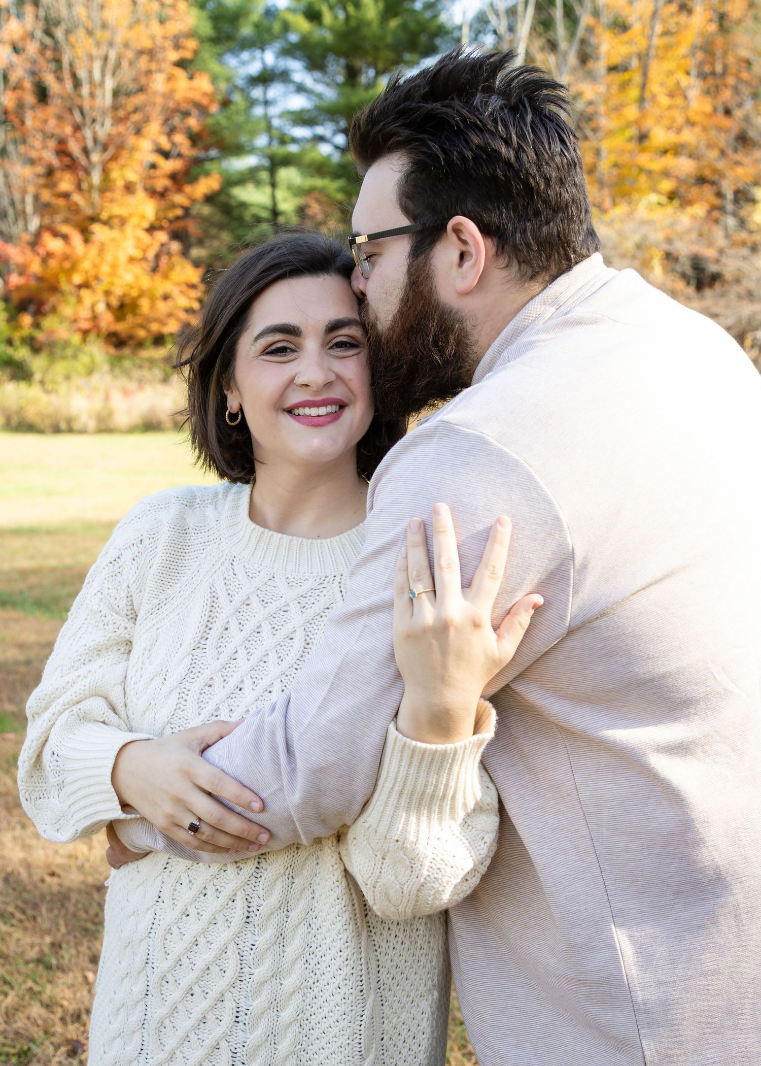 A bearded man kisses the forehead of a smiling woman in a cream cable-knit sweater, showing her ring, against colorful autumn foliage.