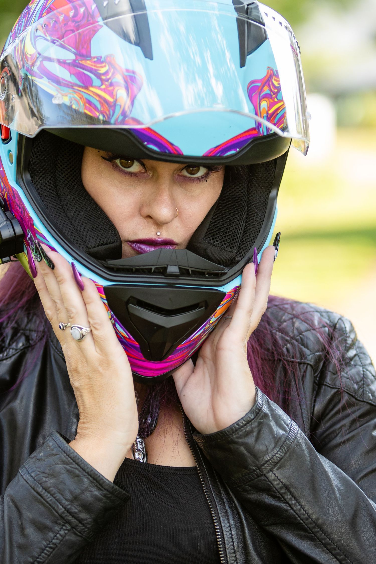 Woman with purple hair and purple lipstick adjusts a colorful motorcycle helmet while wearing a black leather jacket.