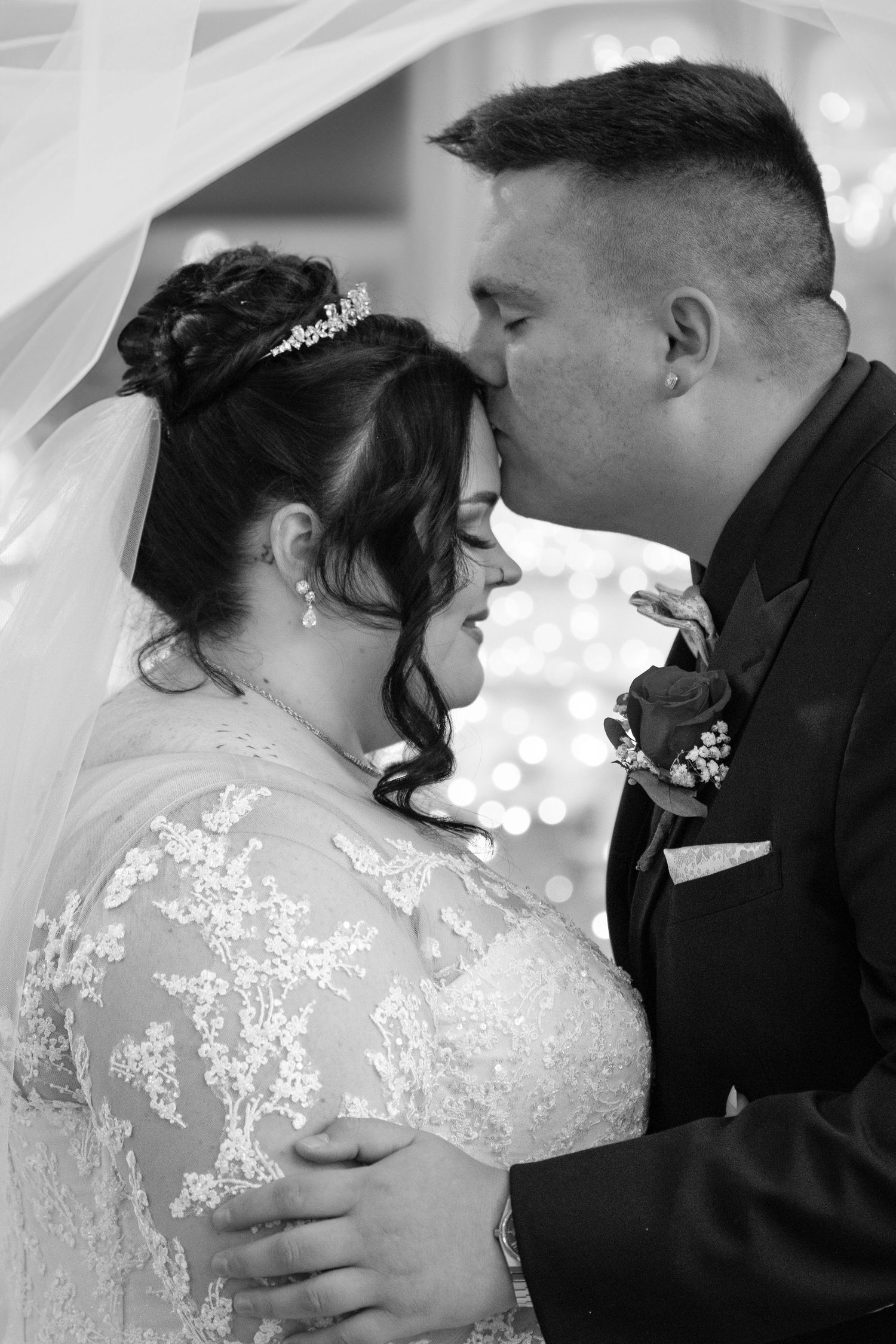 Black and white portrait of a groom tenderly kissing his bride's forehead as she stands in a lace gown and tiara beneath a flowing veil.