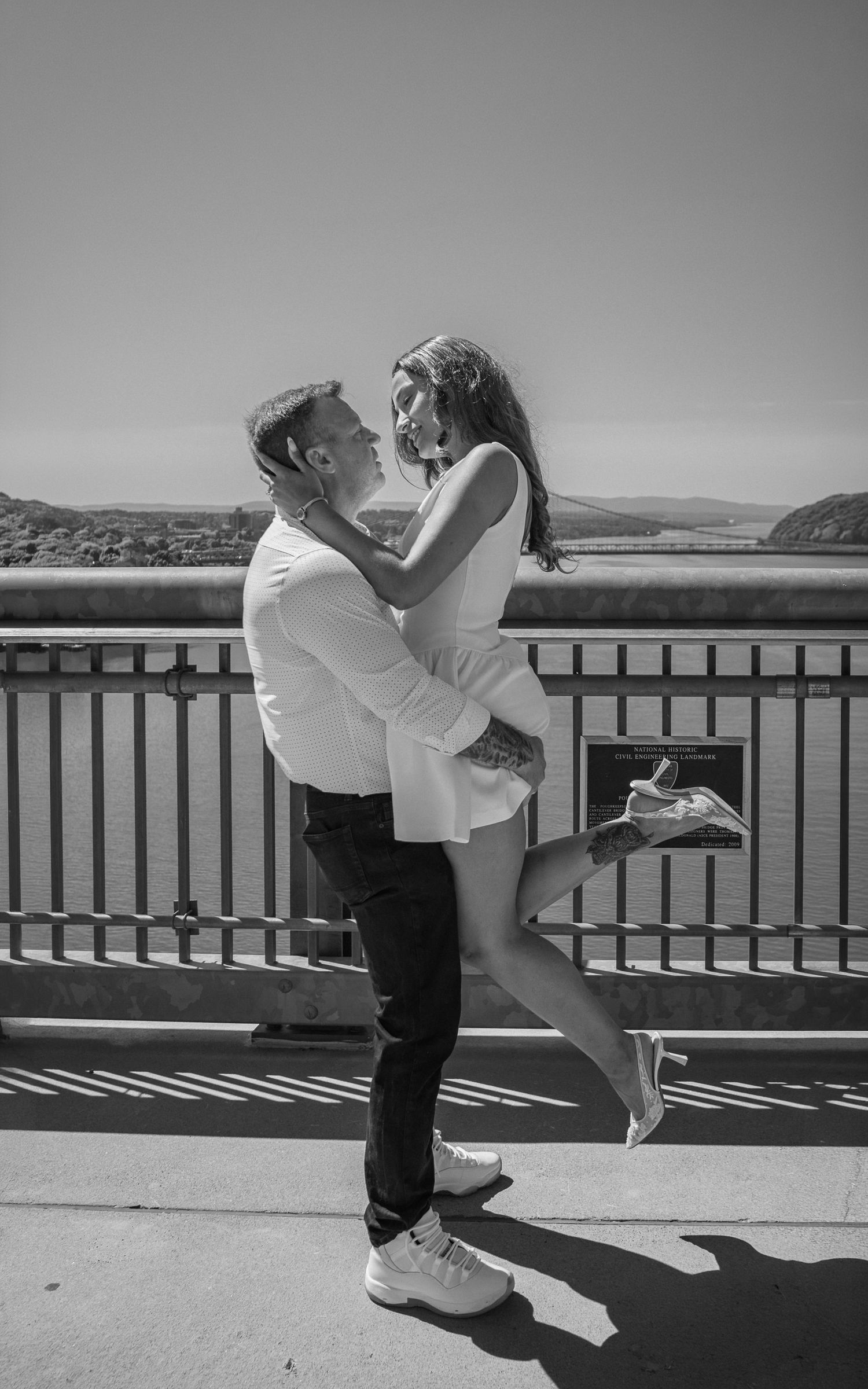 Black and white image of a man lifting a woman in a joyful embrace on a bridge with a scenic river view behind them.