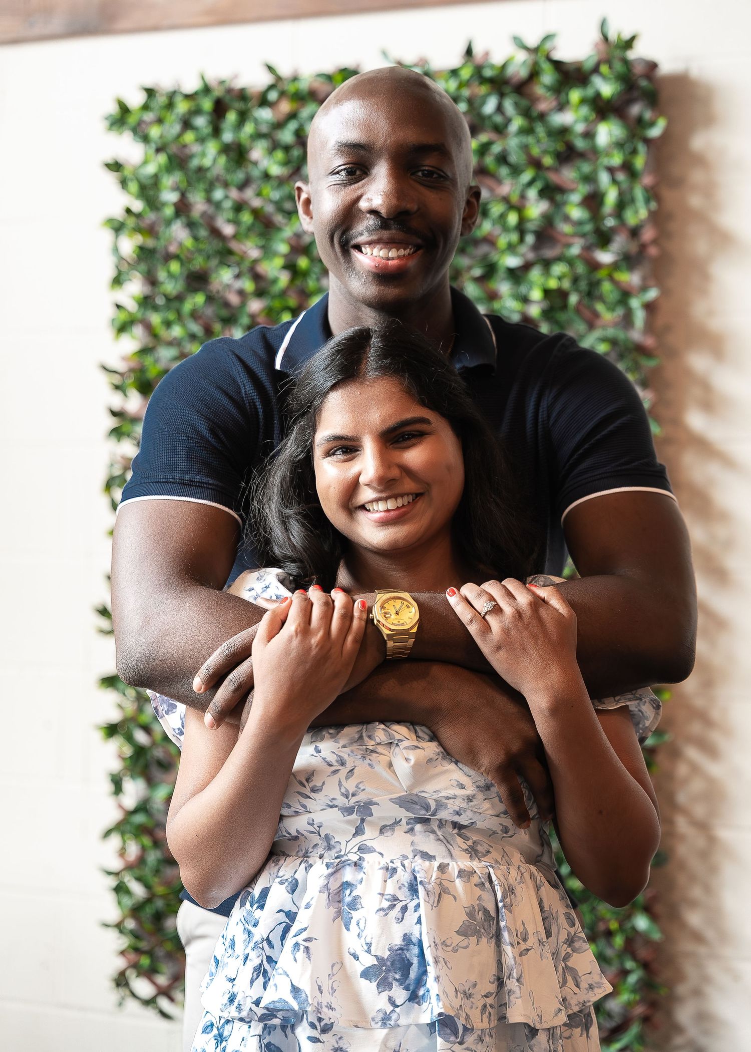 Smiling man in a navy polo embracing a woman in a blue floral dress from behind in front of a green hedge wall.