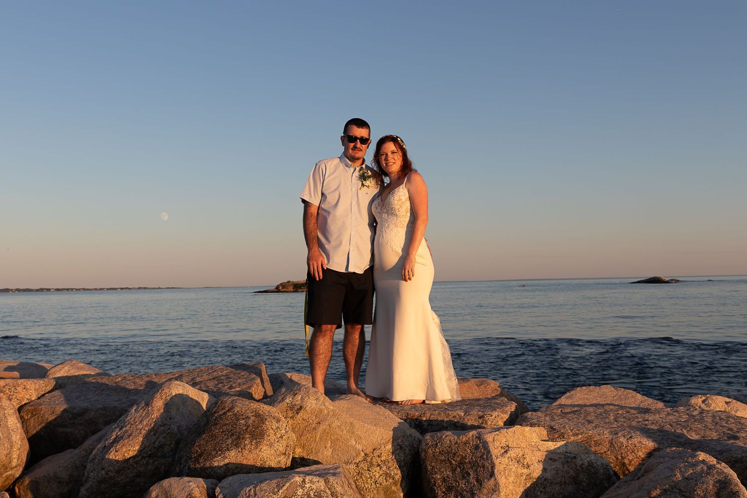 Smiling bride and groom pose together on a rocky jetty beside the calm ocean under a soft sunset sky.