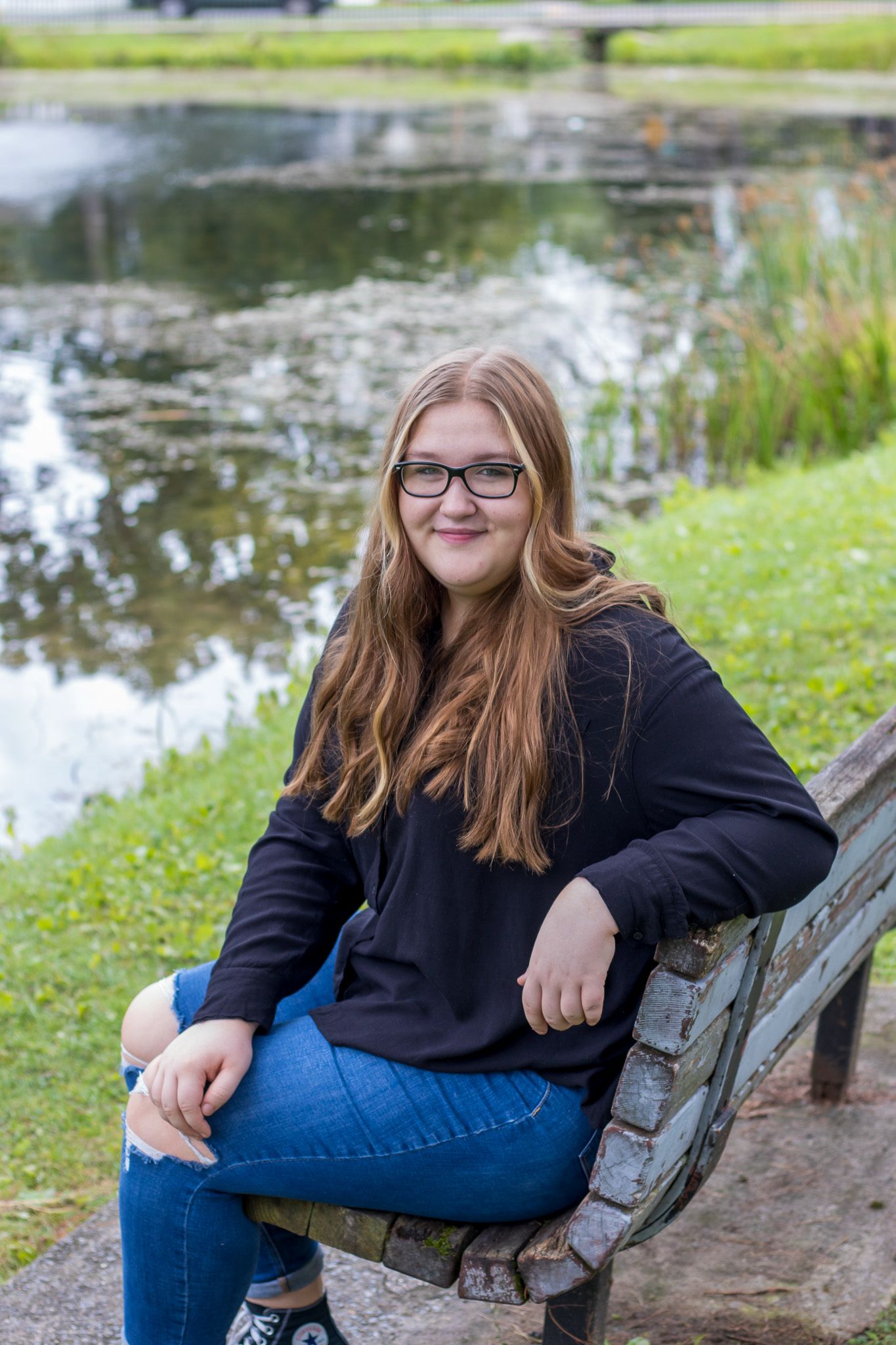 Smiling young woman with long hair and glasses sits on a park bench in a black sweater and jeans beside a pond.