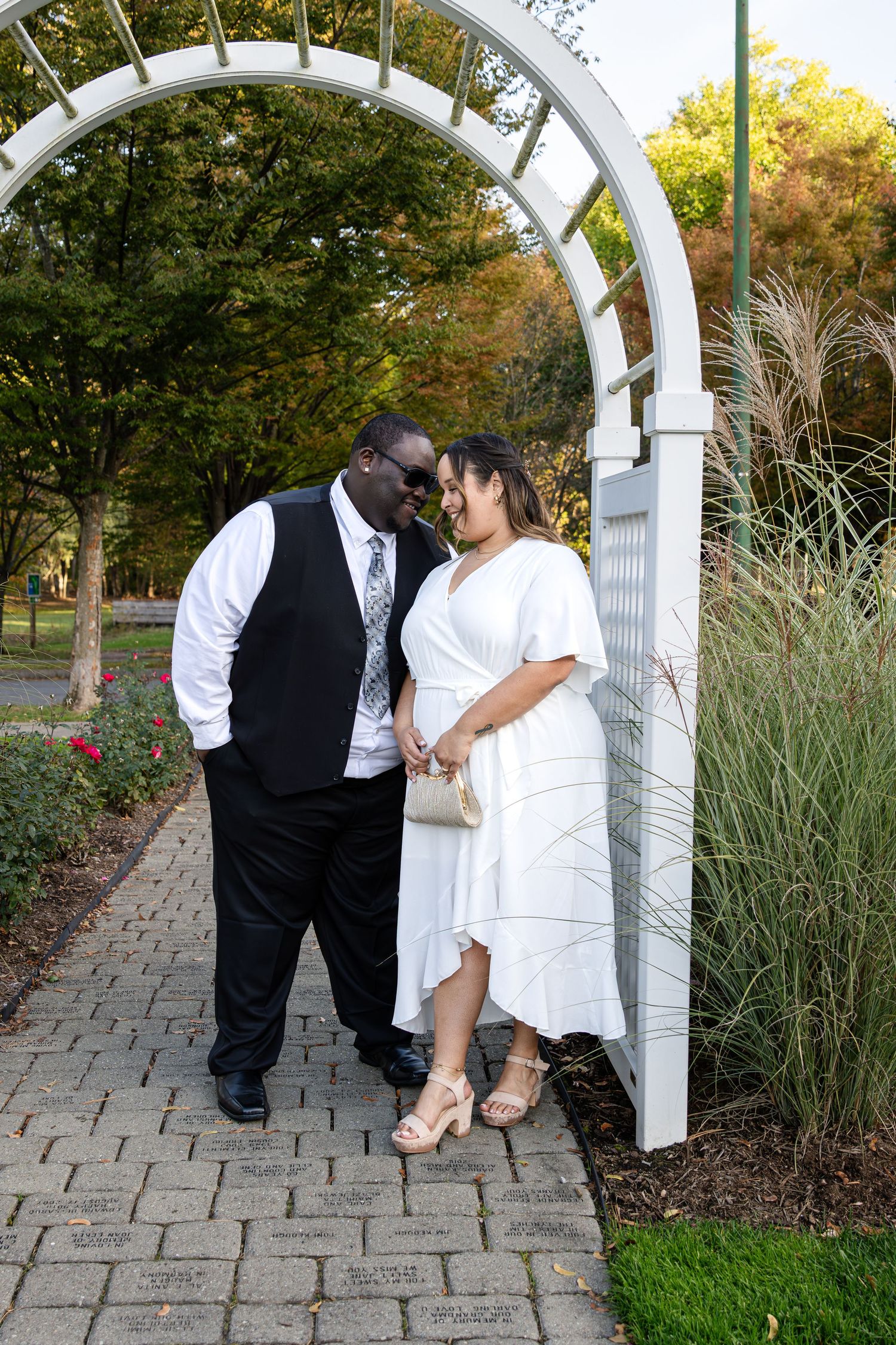 Couple in formal attire share a close moment beneath a white garden arch on a brick path surrounded by autumn trees and tall grass.