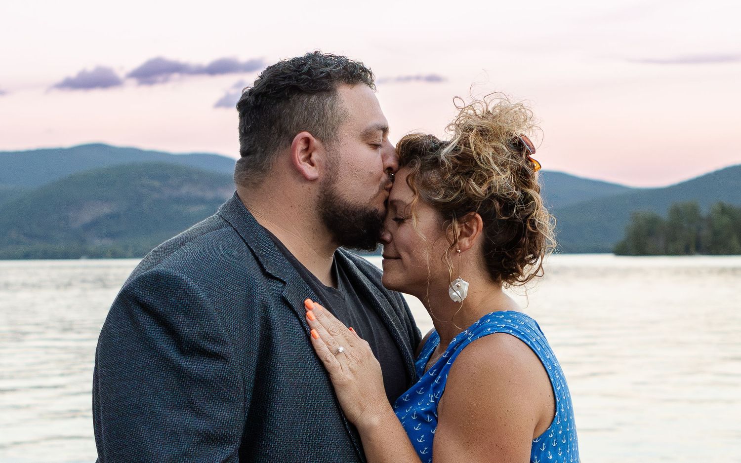 A bearded man in a gray blazer kisses the forehead of a woman in a blue top beside a lake at sunset after an engagement.