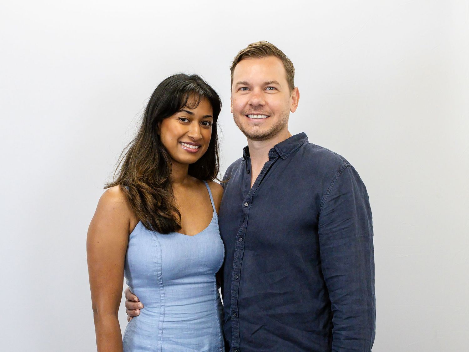 A smiling woman in a light blue dress and a man in a navy shirt pose together against a plain white studio background.