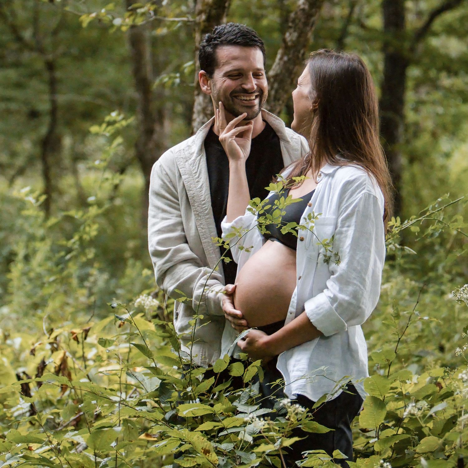 A laughing man gently touches his pregnant partner's belly as they stand among green forest foliage during a maternity photo session.