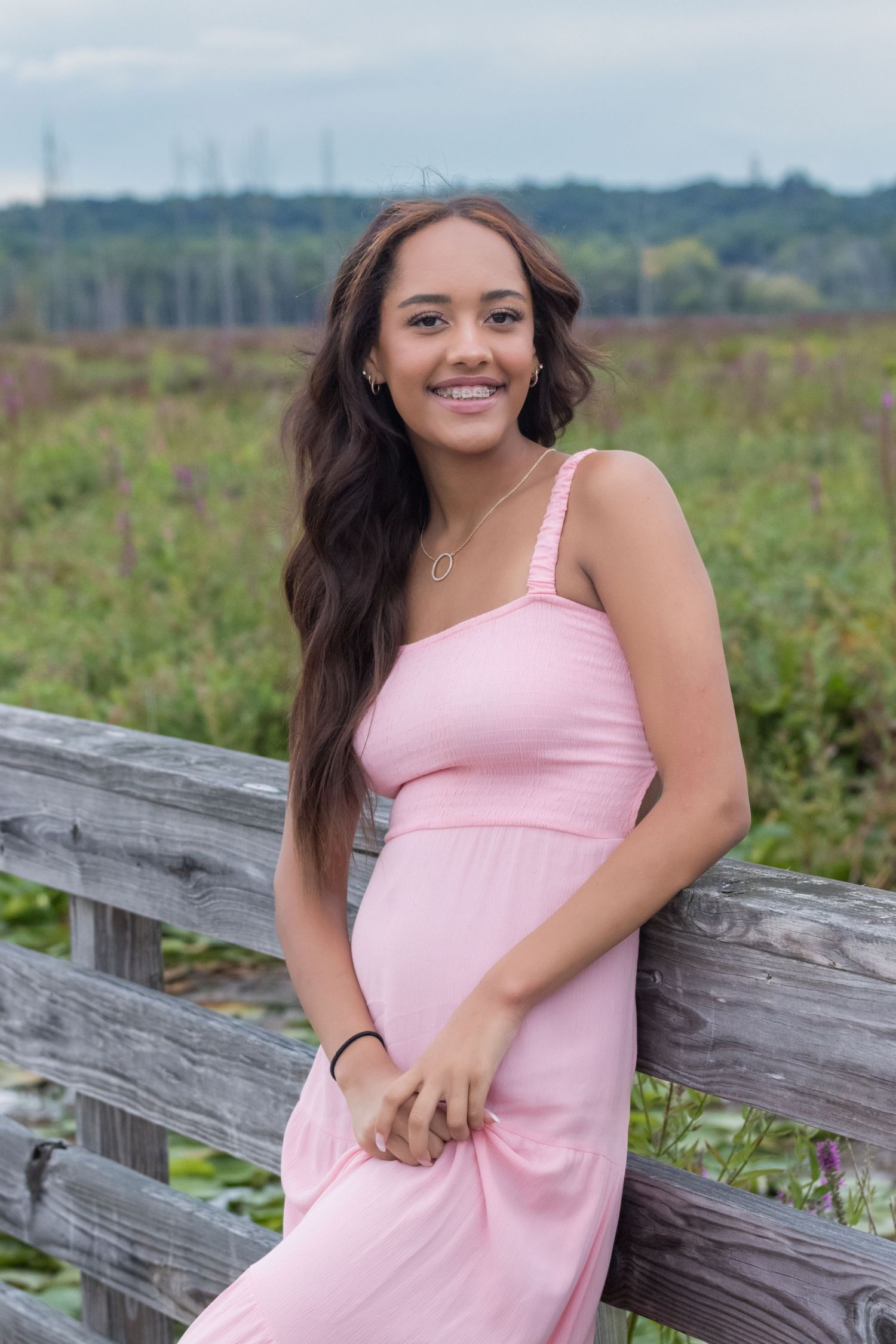 Young woman in a pink sundress smiling as she leans on a weathered wooden railing with a green meadow behind her.