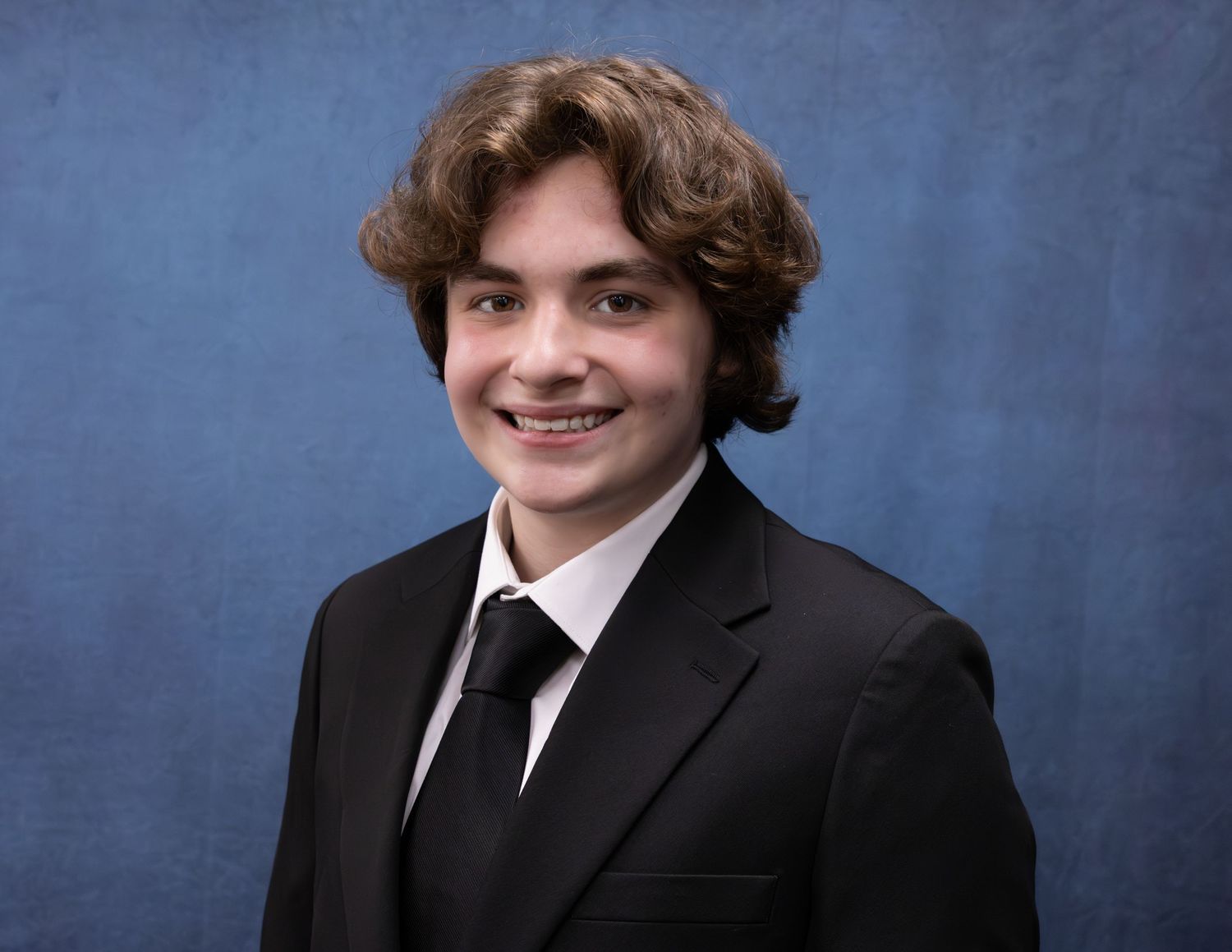 Smiling teenager with curly brown hair in a black suit and tie poses against a blue studio background.