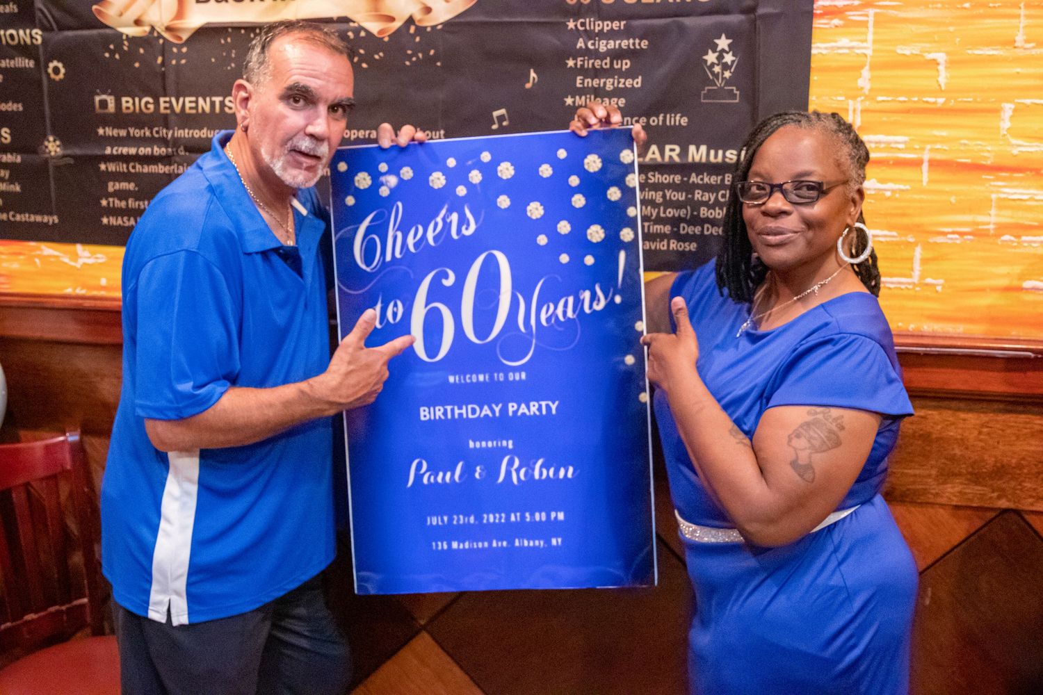 Man and woman in matching blue outfits pose beside a blue Cheers to 60 Years birthday party sign inside a restaurant.
