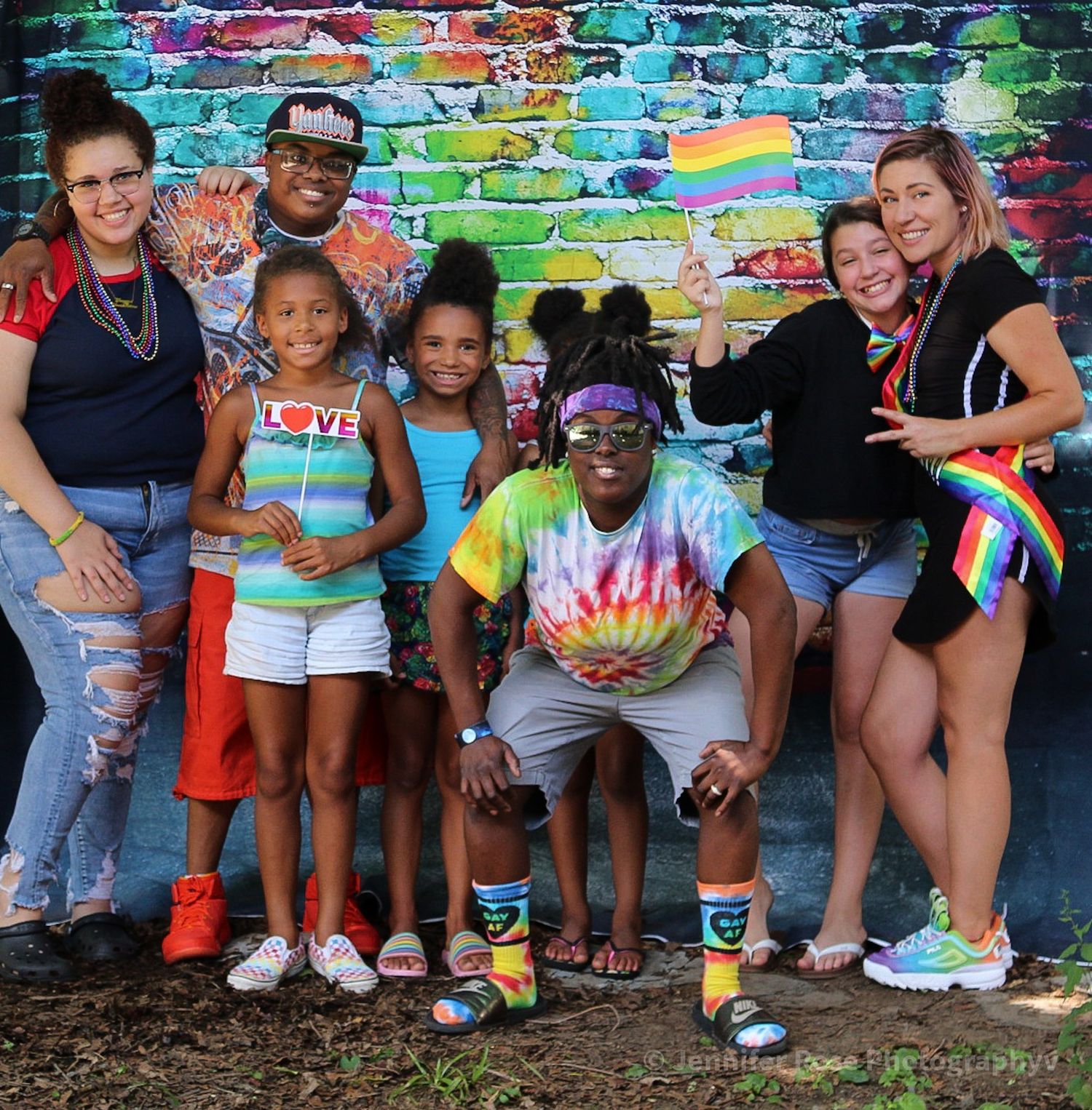 Smiling group of adults and children posing together in front of a colorful brick mural, holding small rainbow pride flags.