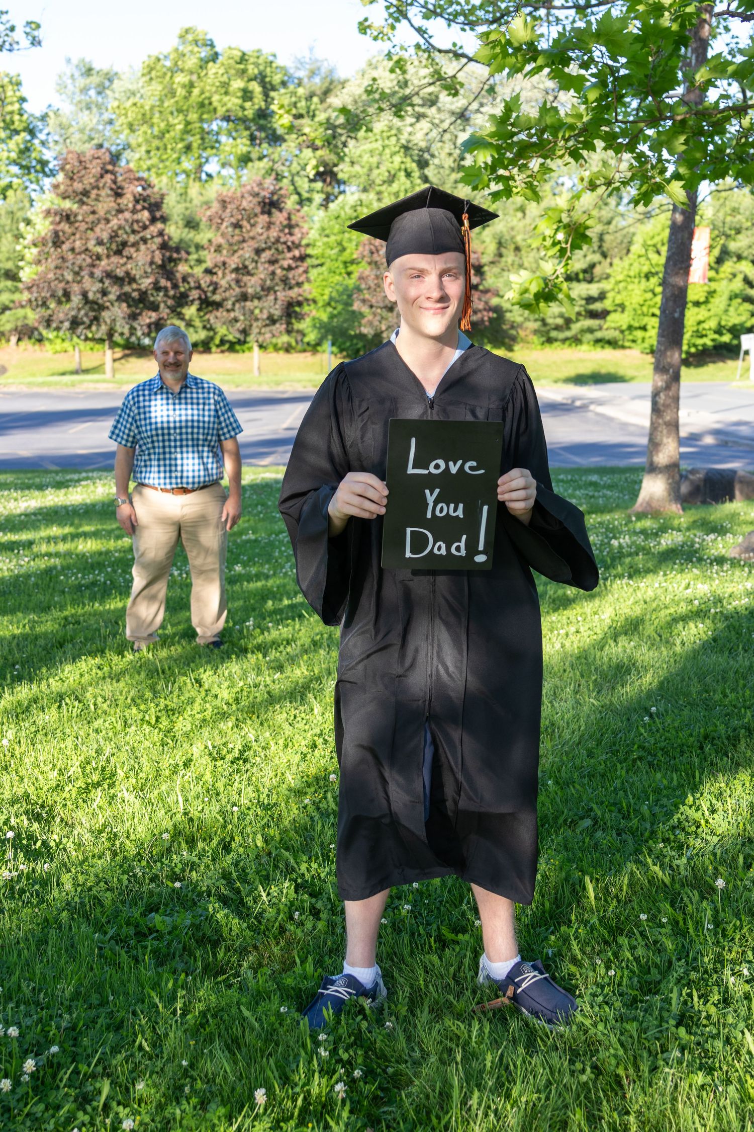 A smiling graduate in a cap and gown holds a chalkboard sign reading Love You Dad while a man stands on the lawn behind him.