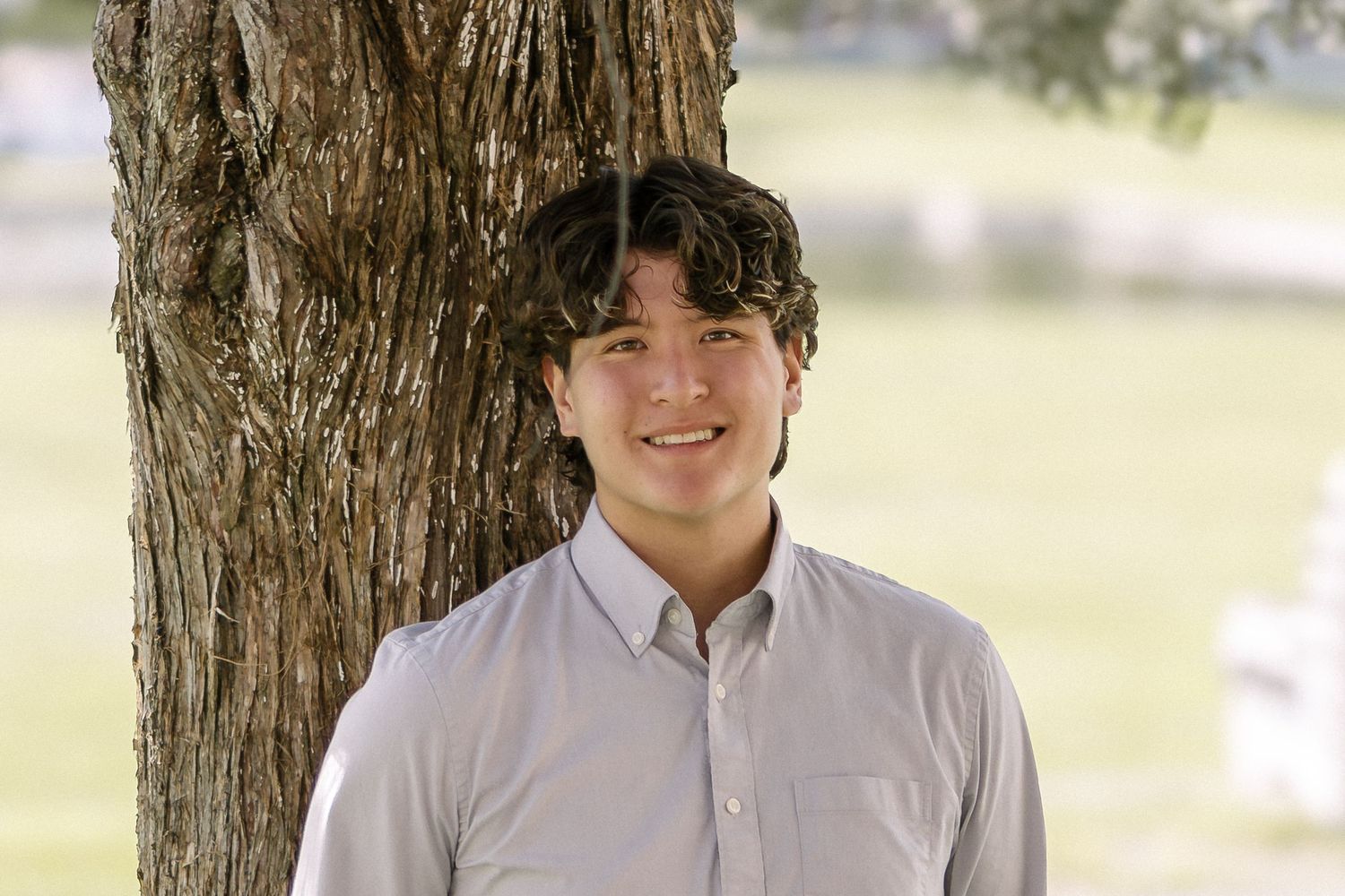 Smiling young man with dark wavy hair in a light gray button-up shirt stands beside a large tree in a sunny park.