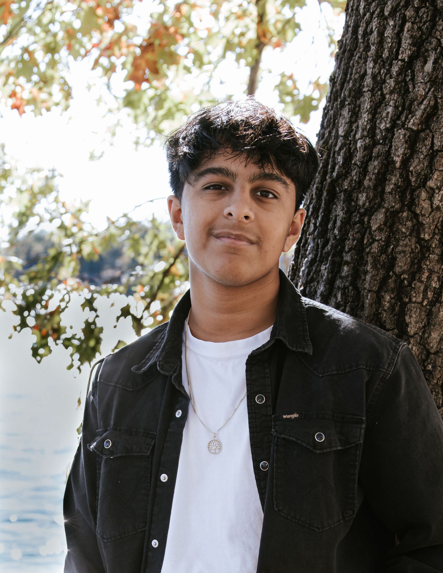 A young man in a black denim jacket and white tee with a pendant necklace stands beside a tree near a sunlit lake.