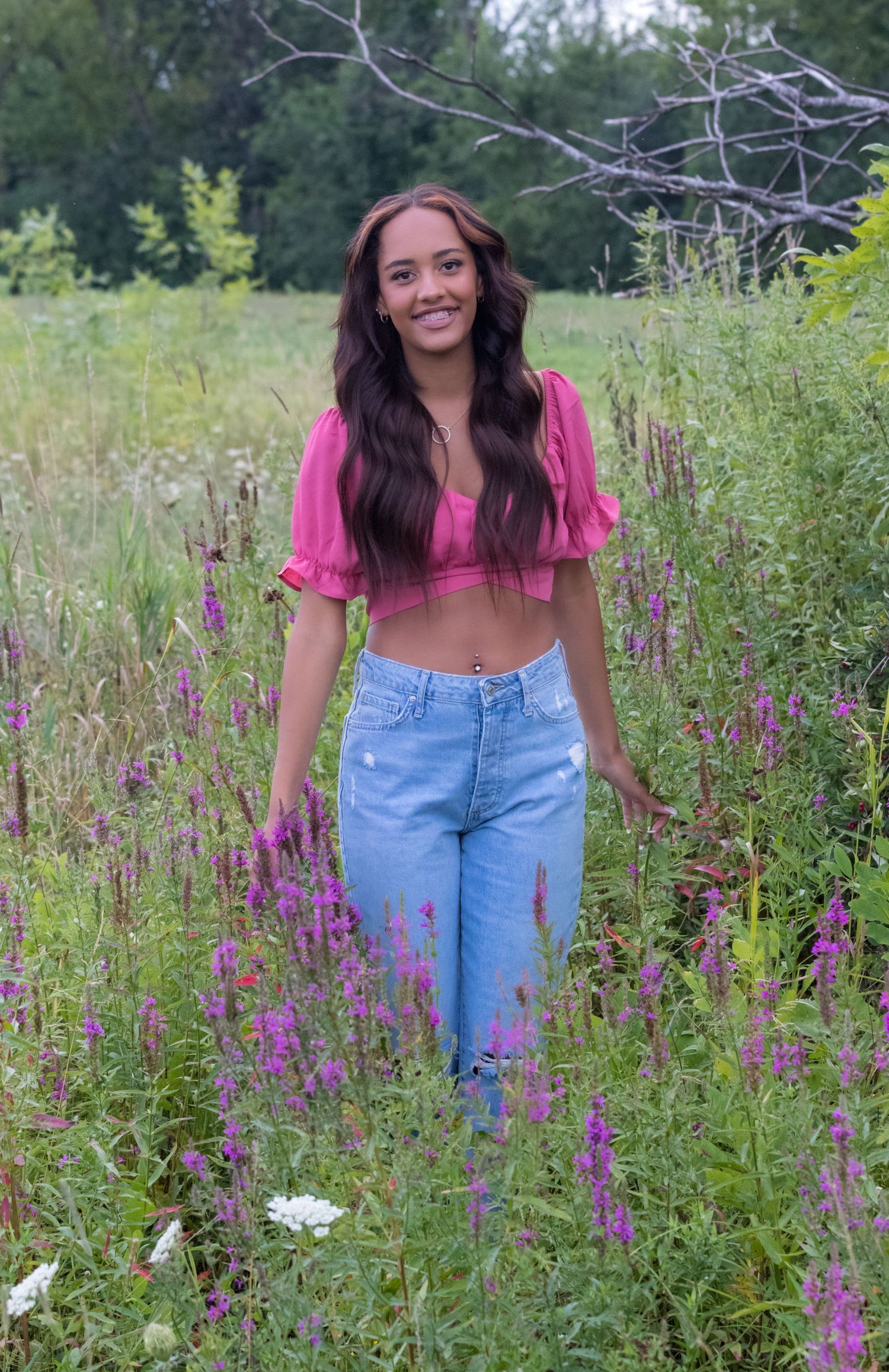 Young woman in a pink crop top and blue jeans walks through a meadow of purple wildflowers on a bright summer day.