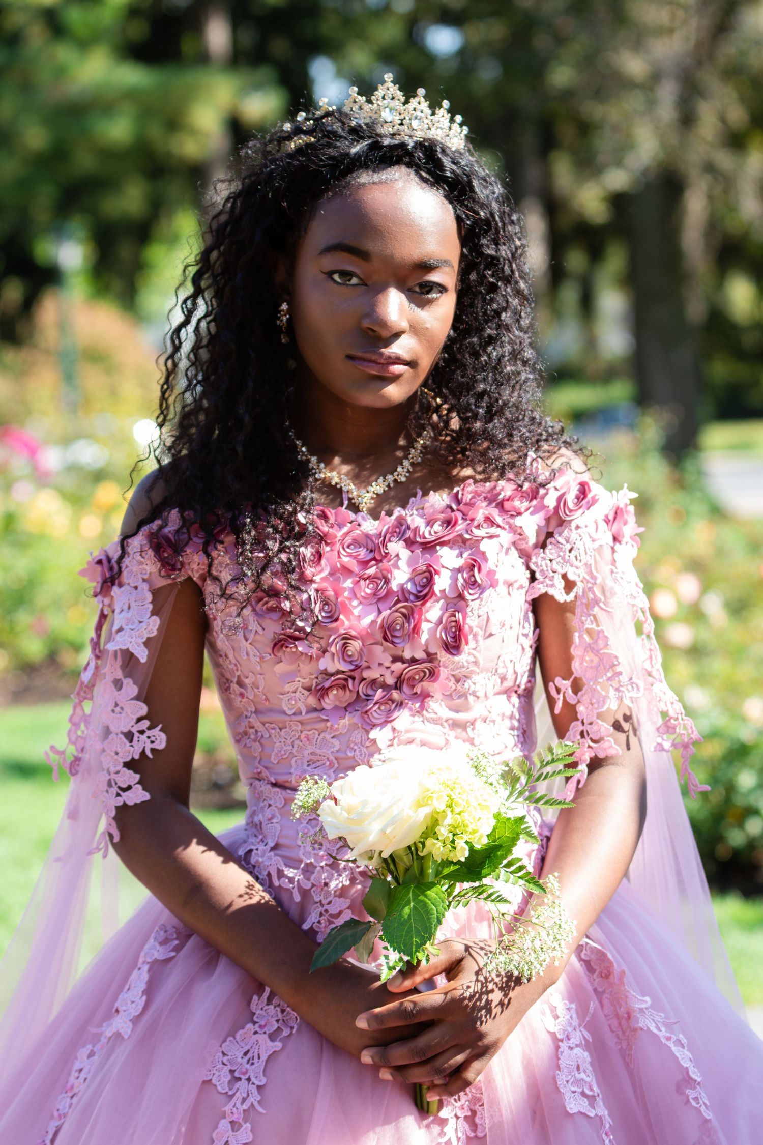 Young woman in a pink embroidered gown and crown holds a white rose bouquet in a sunlit garden.