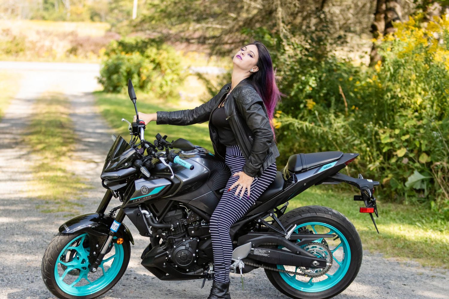 Woman in a leather jacket laughs while sitting on a black motorcycle with teal wheels on a country road.