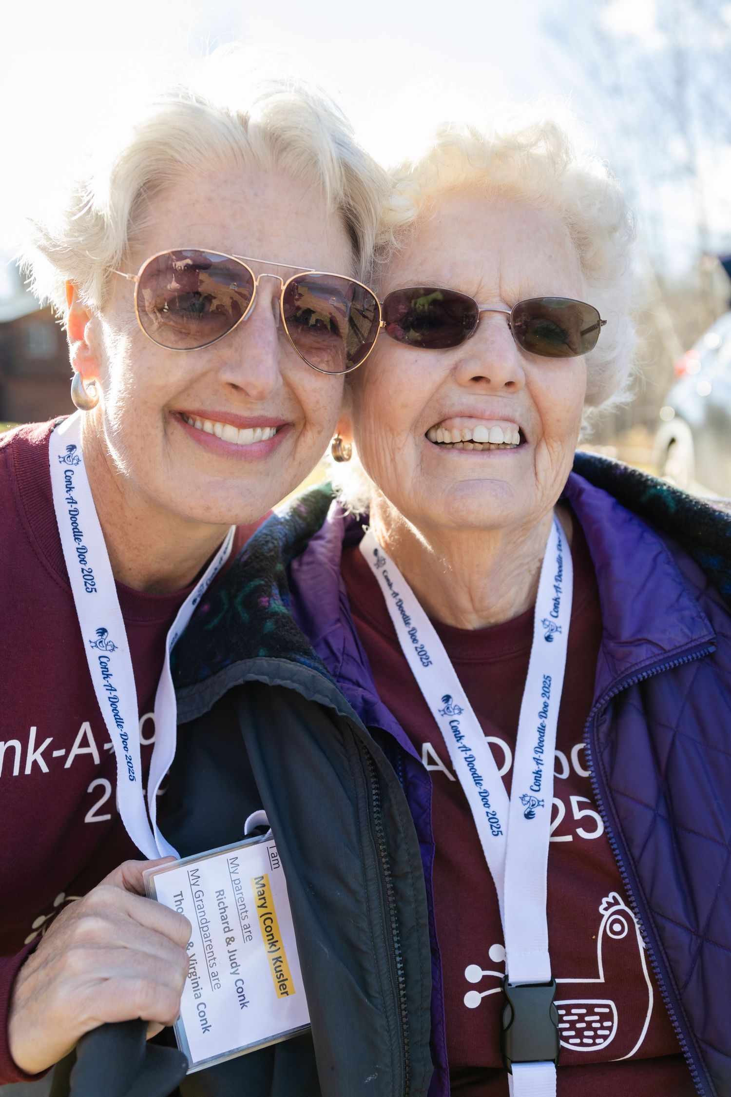 Two smiling women in sunglasses and lanyards wearing maroon shirts pose cheerfully close together outdoors.