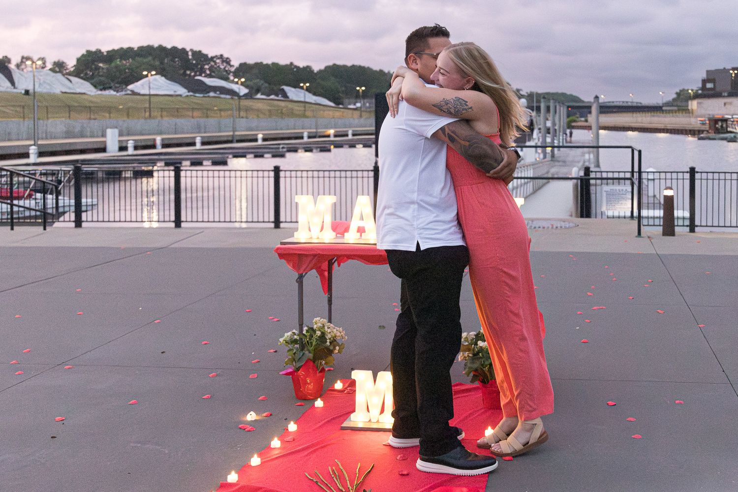 A newly engaged couple embraces on a waterfront walkway beside lit marquee letters, rose petals and candles at dusk.