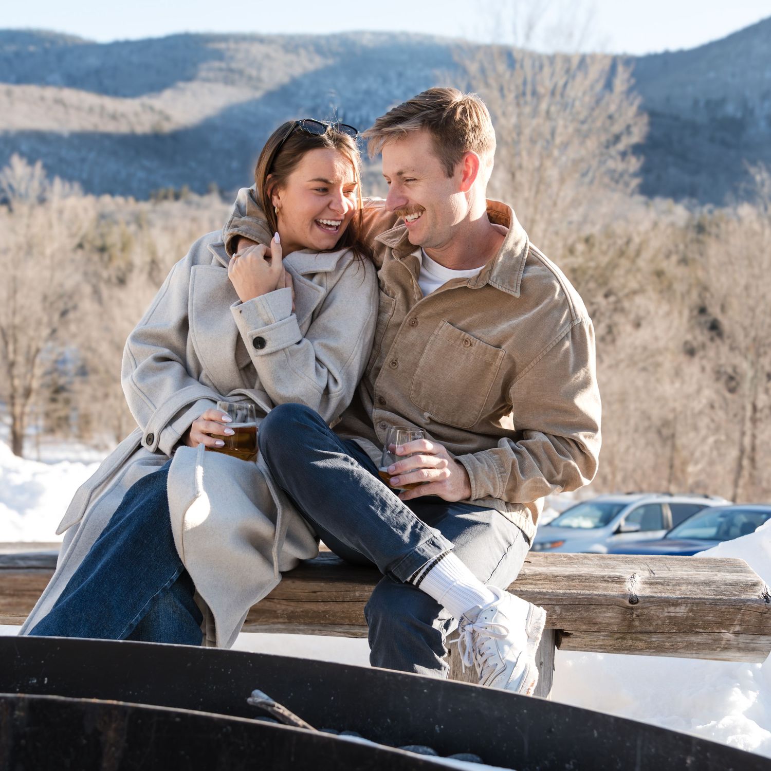 An engaged couple sits close together on a wooden bench, smiling and sharing a warm moment with snowy mountains behind them.