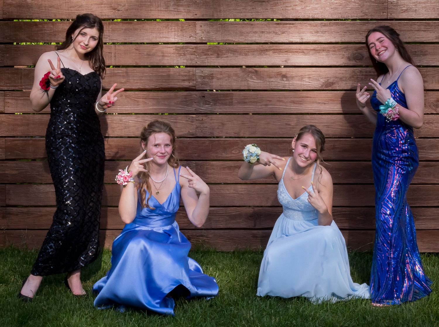 Four young women in colorful prom gowns pose playfully with wrist corsages in front of a wooden fence on a green lawn.