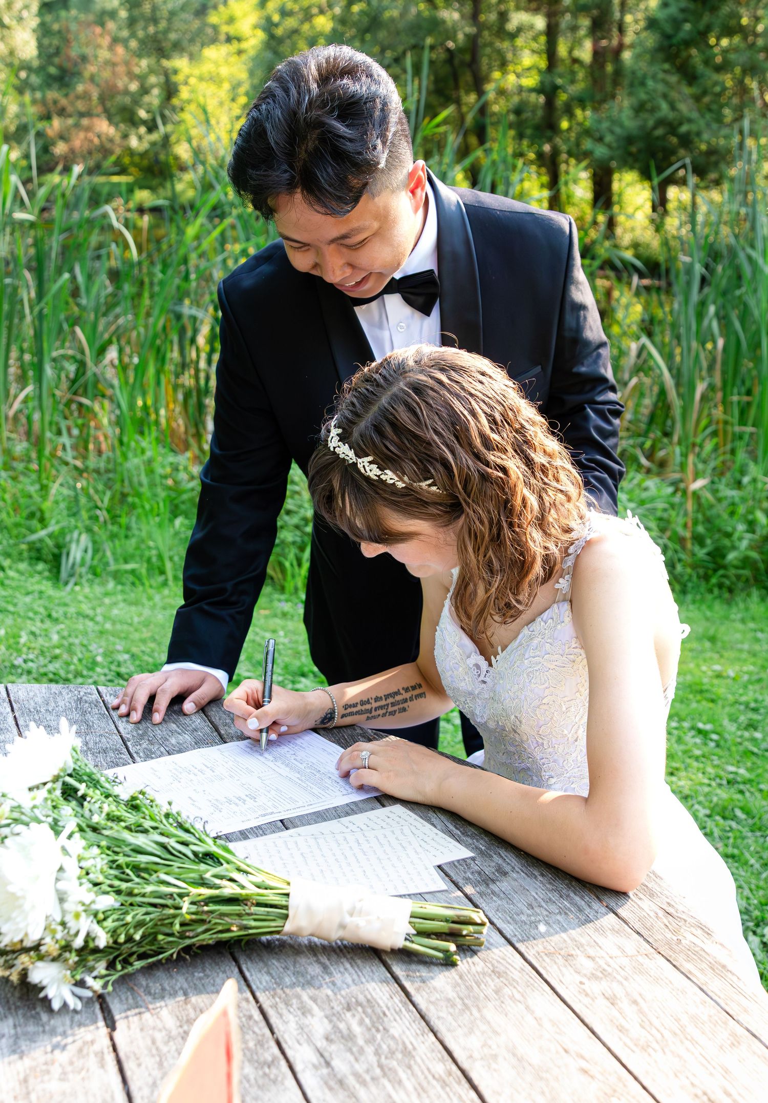Bride in a lace gown and floral headband signs the marriage license at a wooden table outdoors while the groom in a tuxedo looks on.