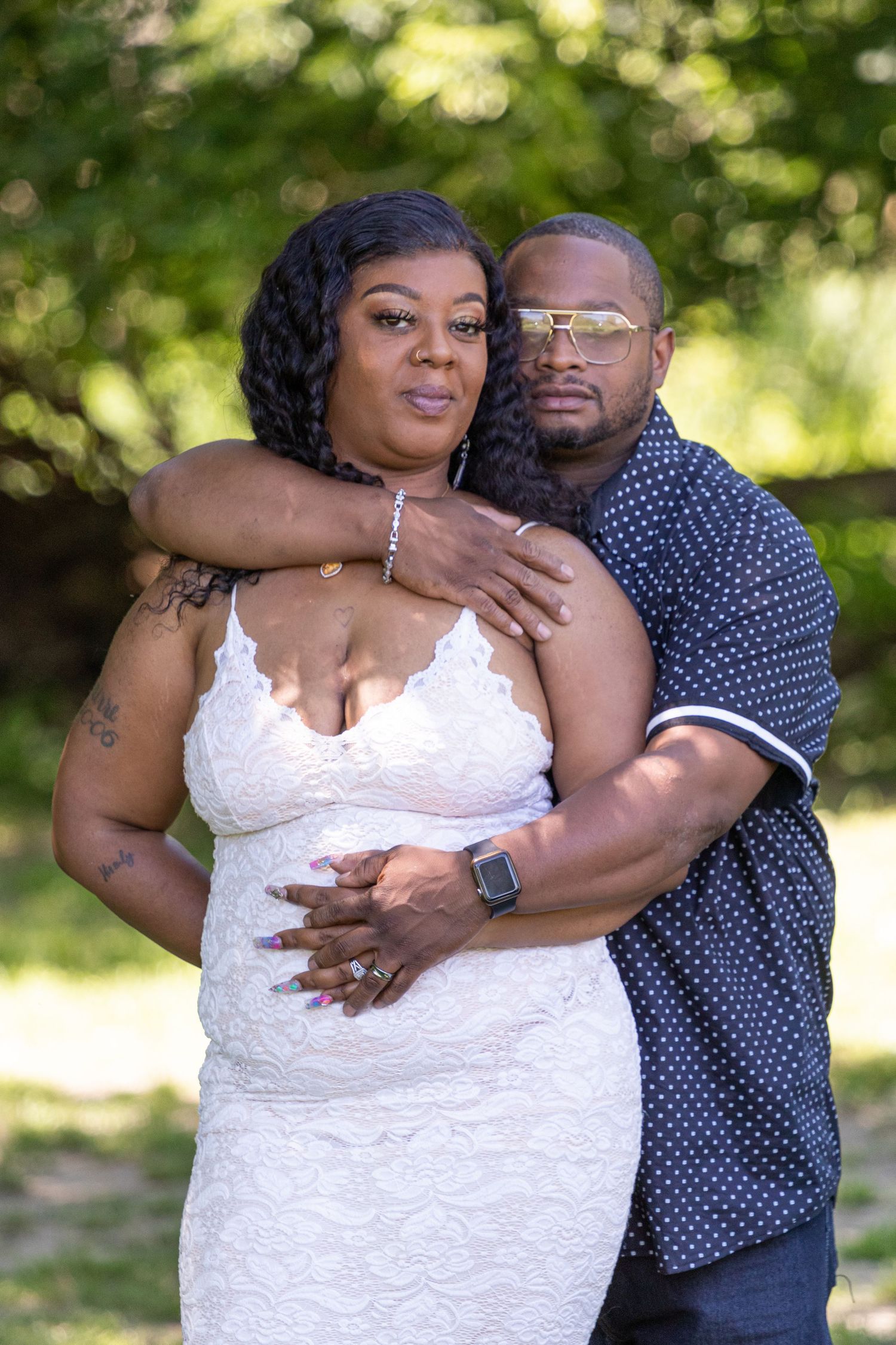 Smiling couple embraces outdoors as a man in sunglasses wraps his arms around a woman in a white lace dress.