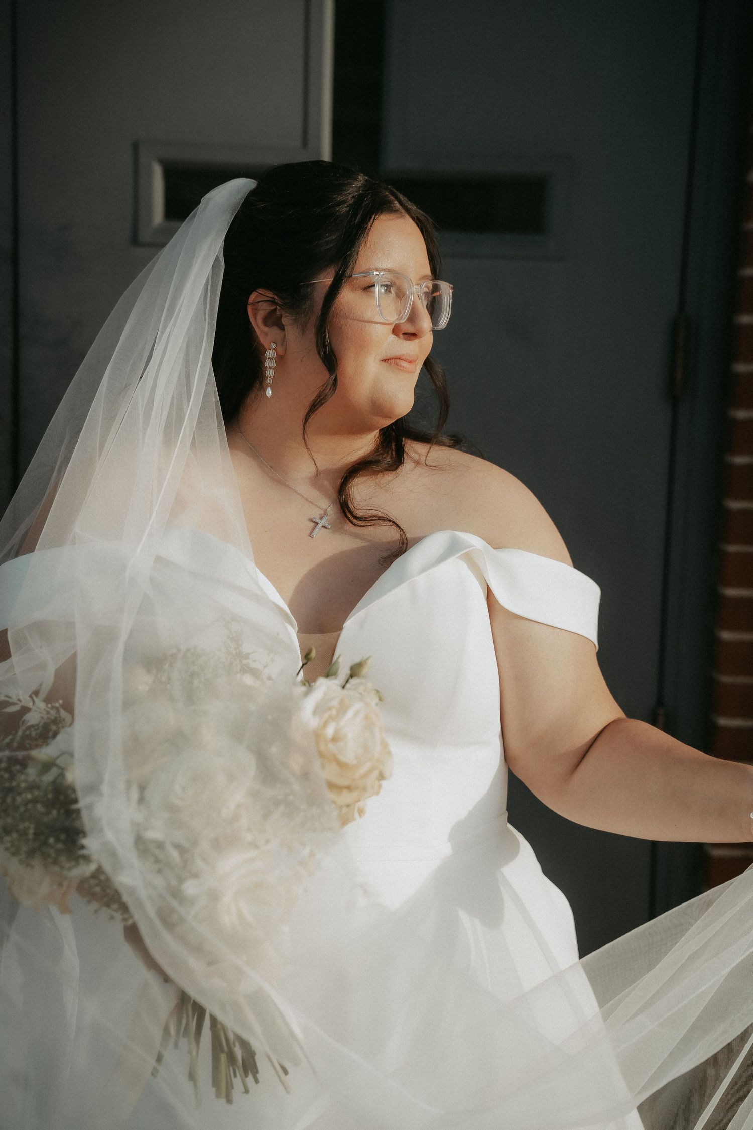 A bride in a white off-shoulder wedding gown and veil poses while wearing glasses in a moody indoor setting.