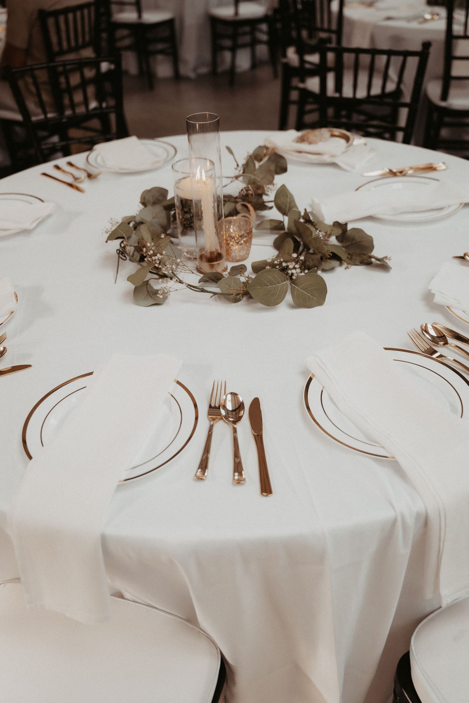 Elegant wedding table setting with white linens, eucalyptus centerpiece, and rose gold place settings.
