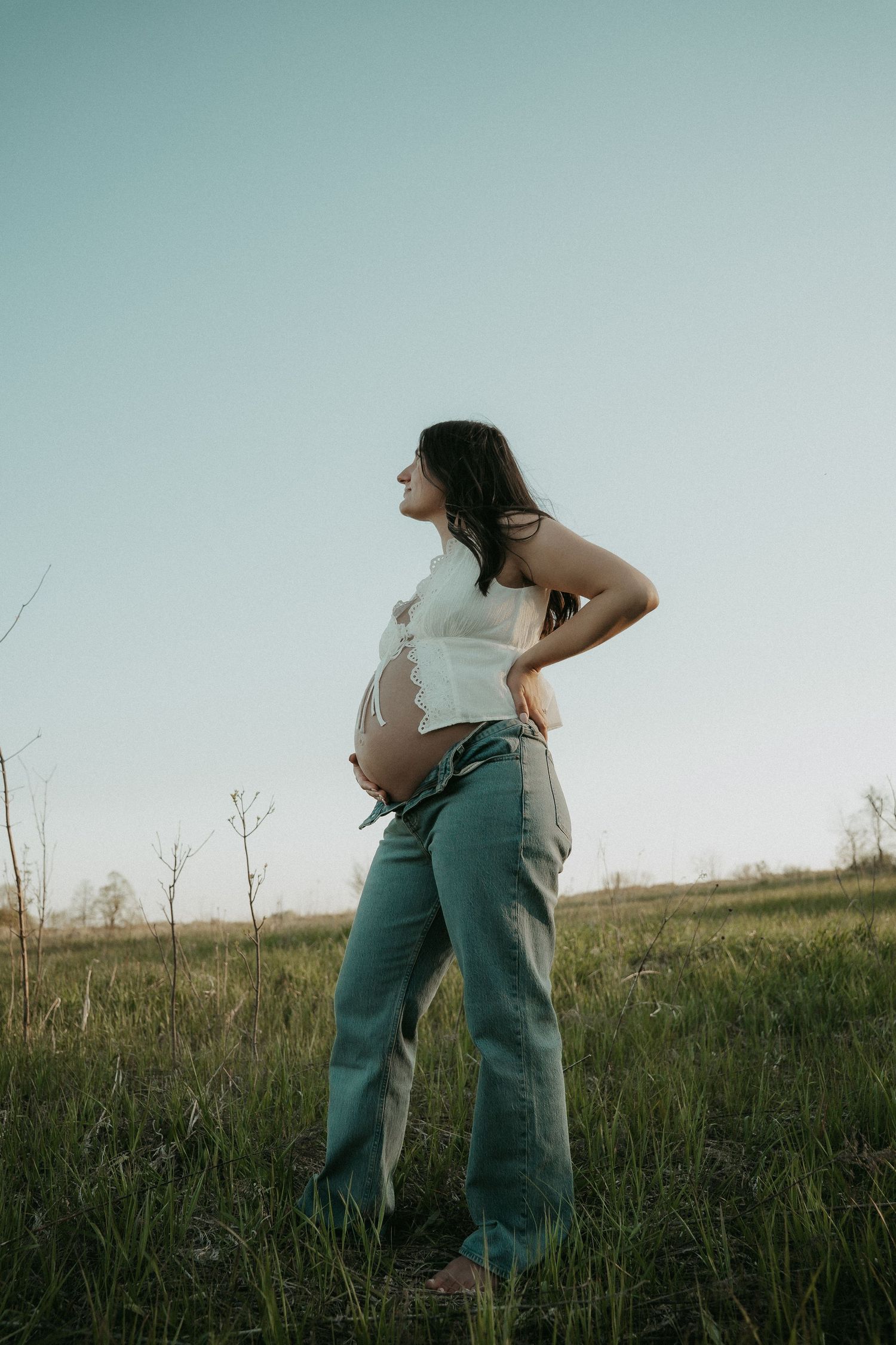 Pregnant woman in white crop top and jeans standing in a green field, cradling her baby bump at sunset.