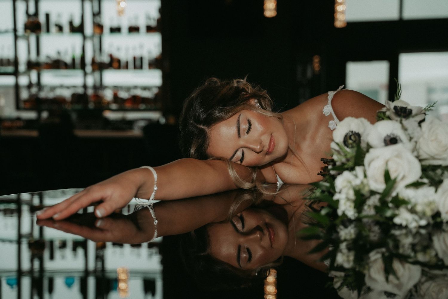 A person rests their head on a reflective surface next to white floral arrangements in a dimly lit setting.