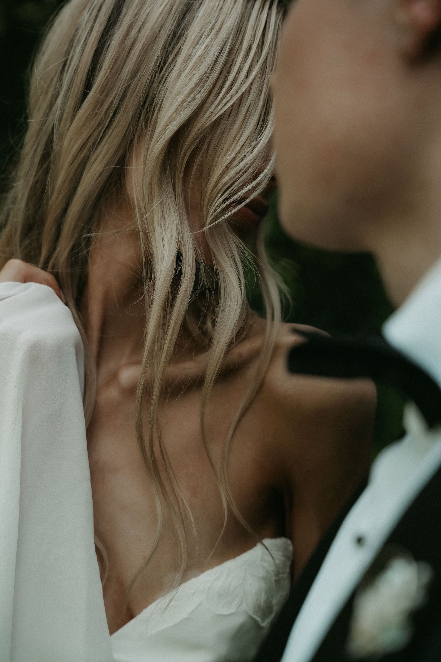 Close-up of a bride with flowing blonde hair in a white off-shoulder gown beside a groom in a black tuxedo.