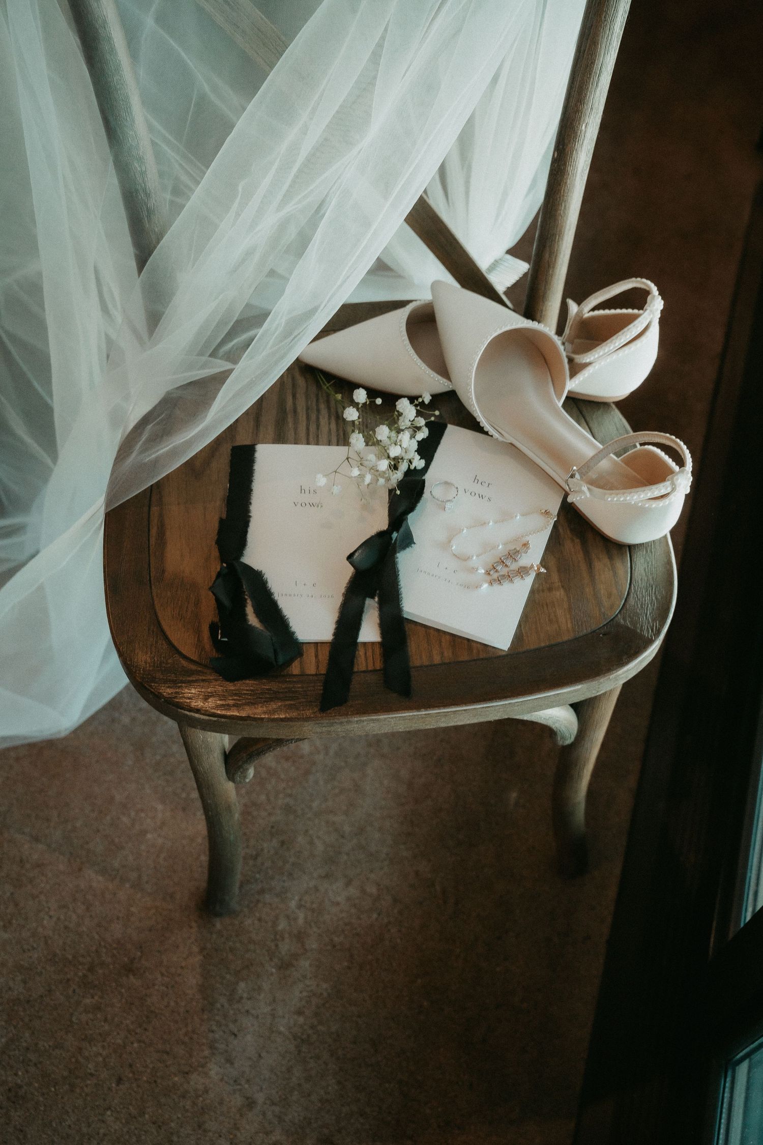 Wedding accessories including high heels, veil and invitation arranged on vintage wooden chair in moody lighting.