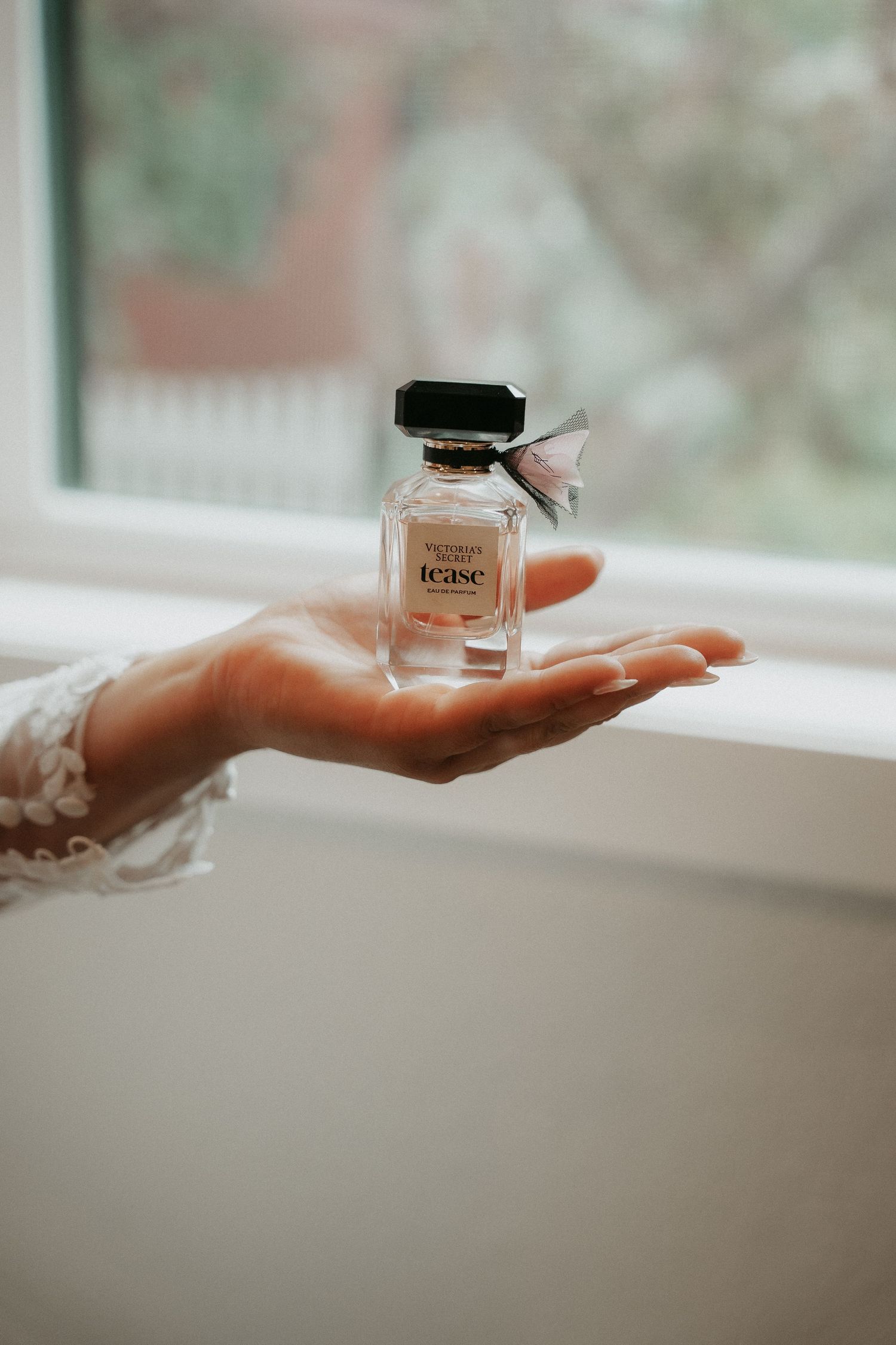 A series of hands holding a small glass perfume bottle against a soft white windowsill background.