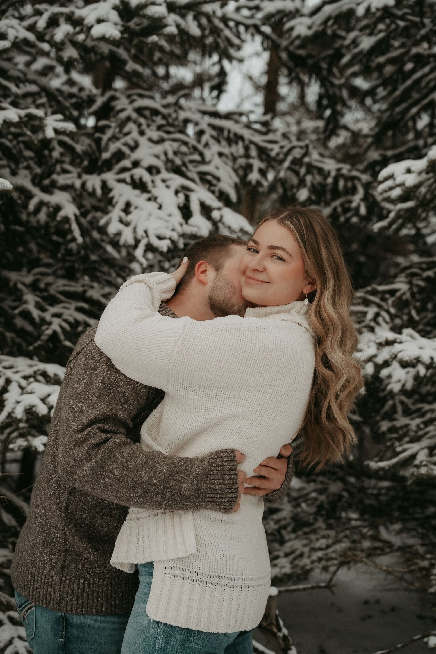 A couple embraces in a romantic winter scene with snow-covered trees in the background.