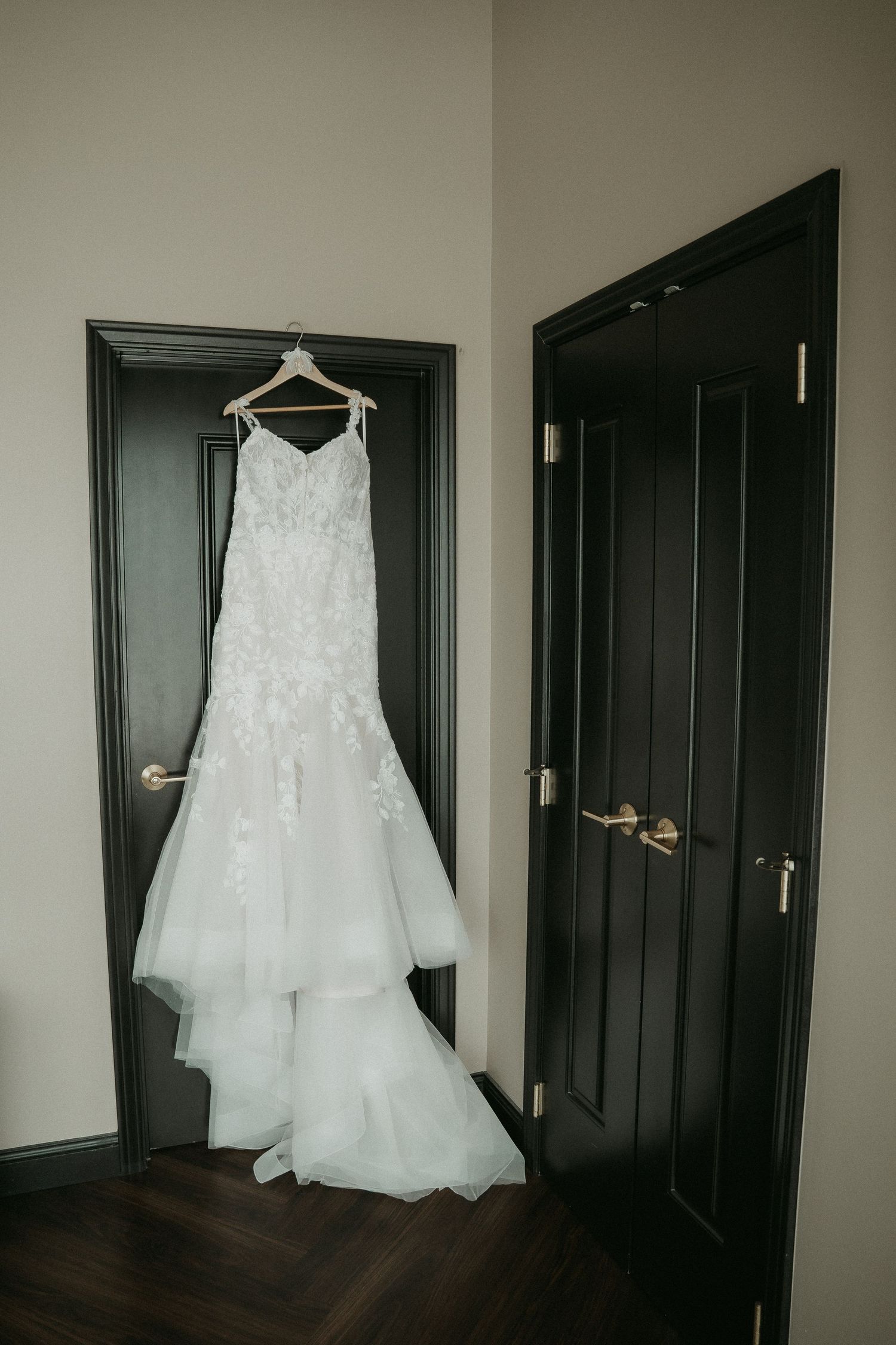A white wedding dress hangs elegantly against a dark wooden door in a dimly lit hallway.