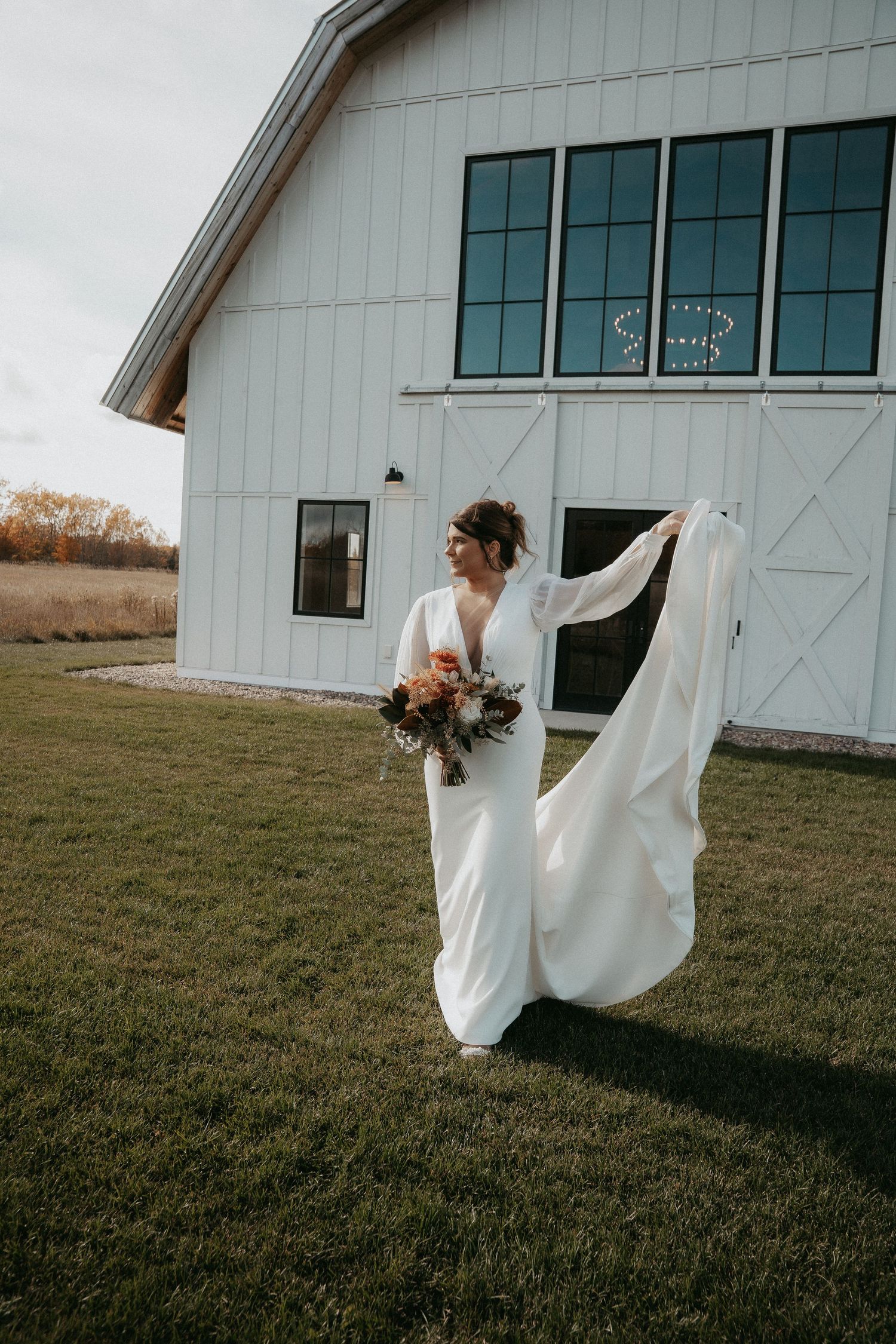 A flowing white wedding dress billows in the wind against a rustic white barn backdrop in a pastoral setting.