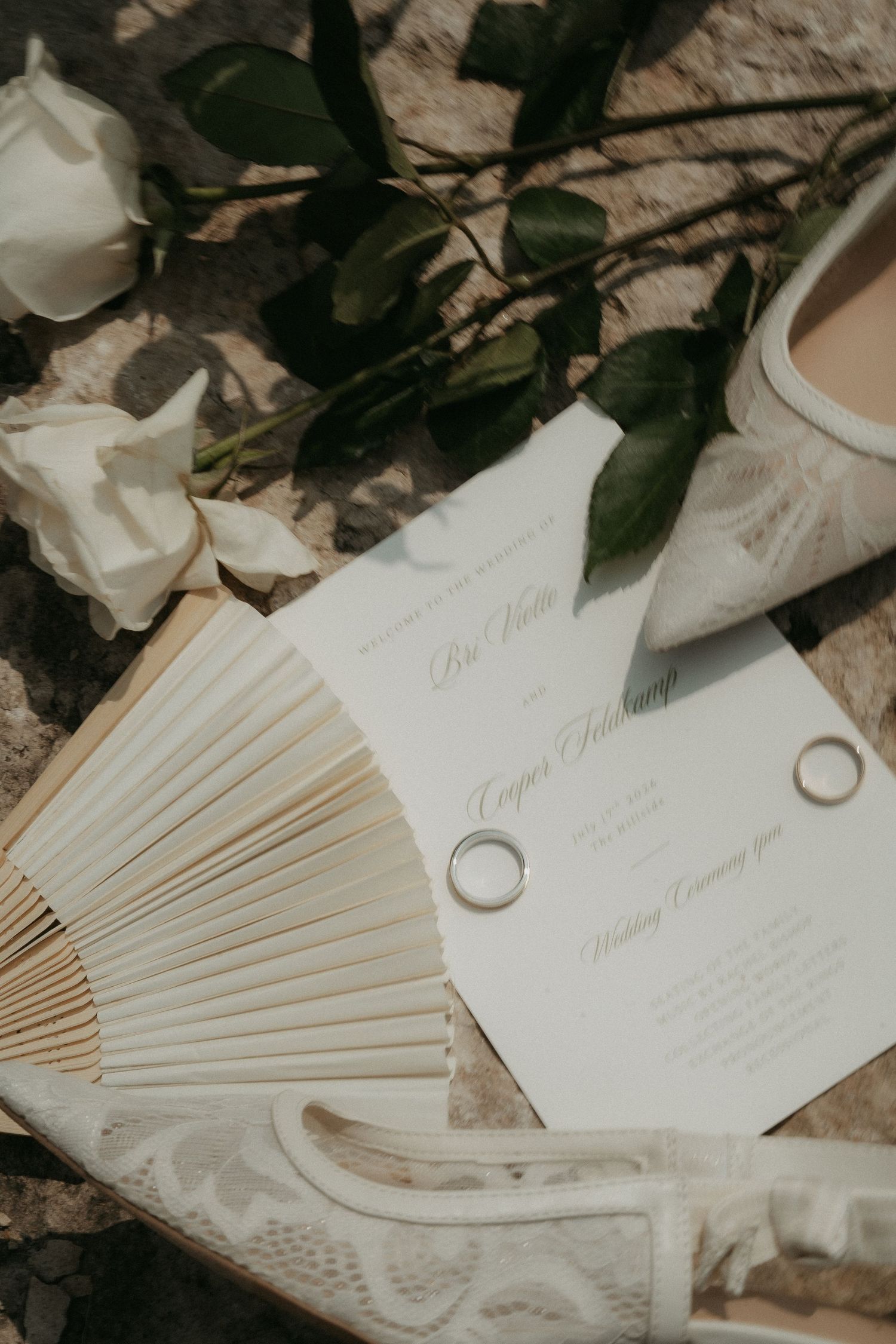 Elegant wedding flat lay with ivory invitation, gold rings, white roses, a paper fan, and lace bridal shoe on stone surface.