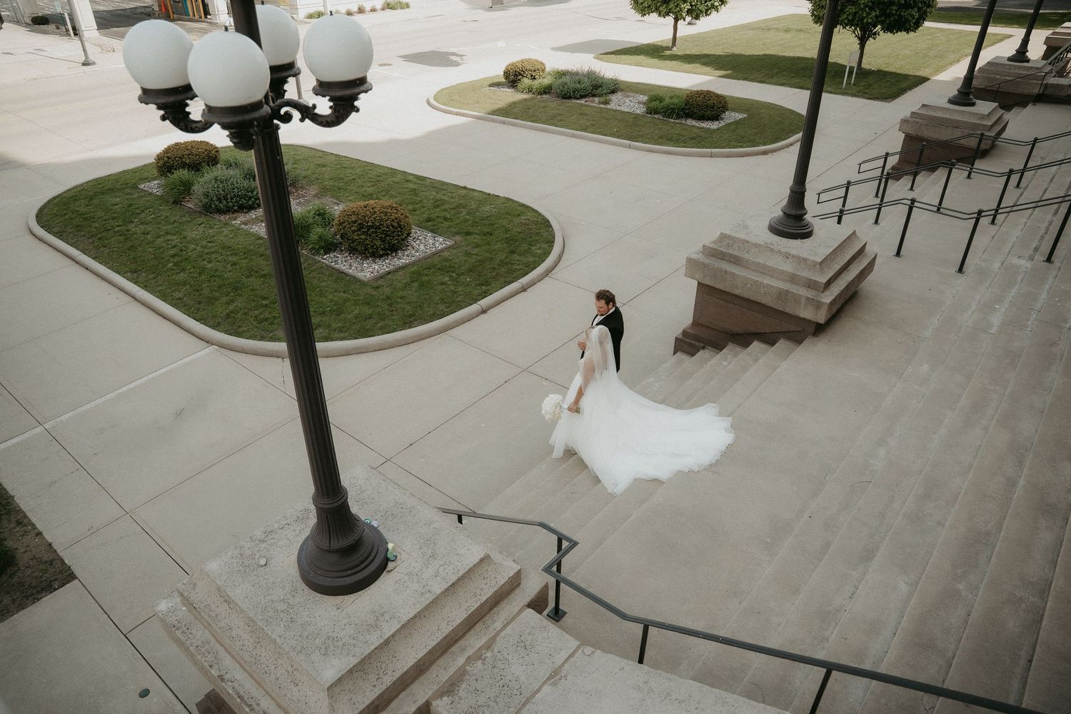 Aerial view of a bride in a flowing white dress walking down concrete steps at an outdoor venue.