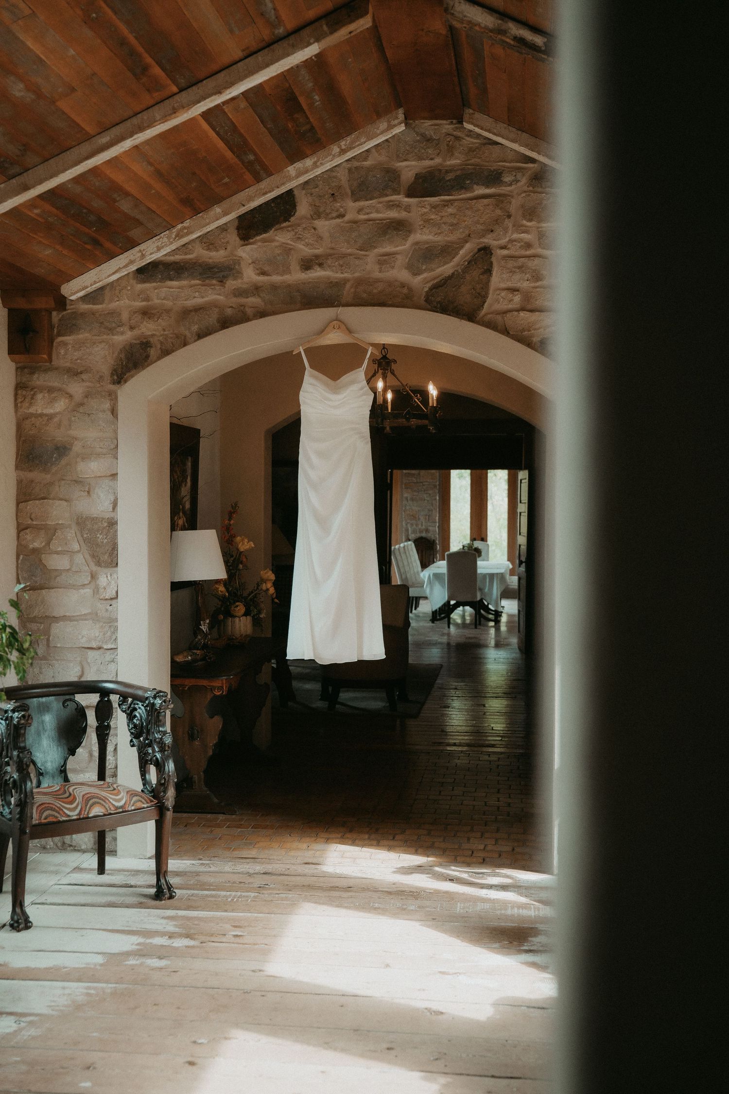 Wedding dresses hang in a rustic stone room with wooden beams and natural light streaming in.